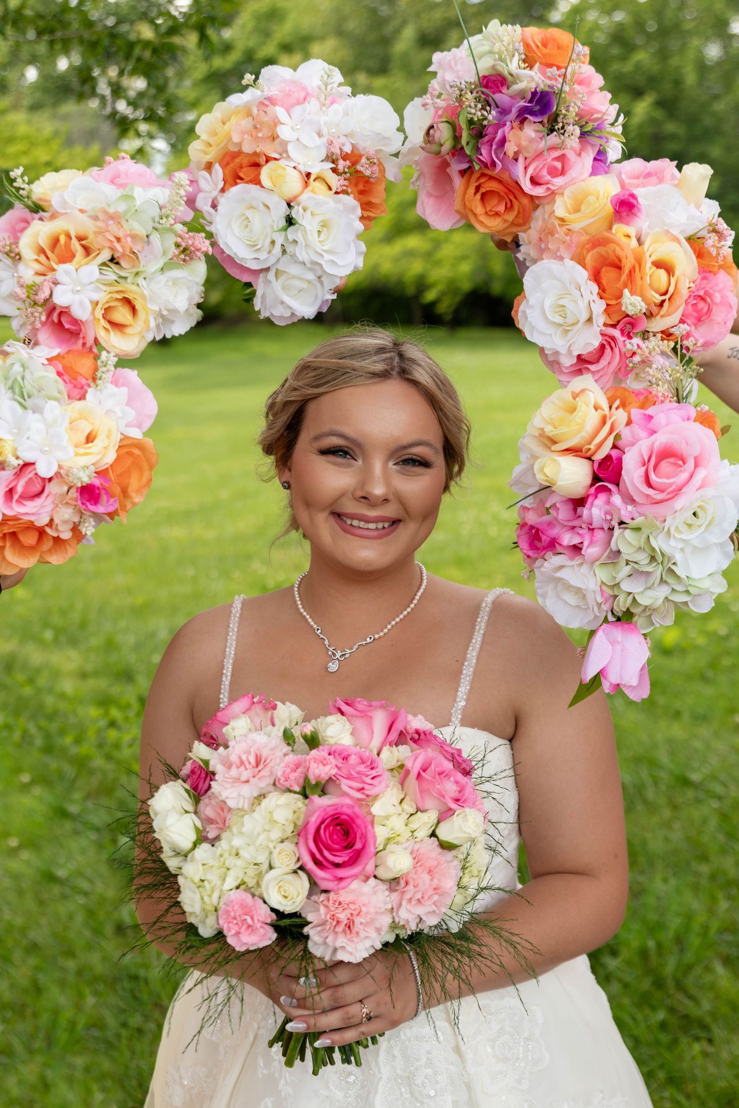 Radiant bride holding pink and white bouquet framed by a colorful floral heart arch outdoors.