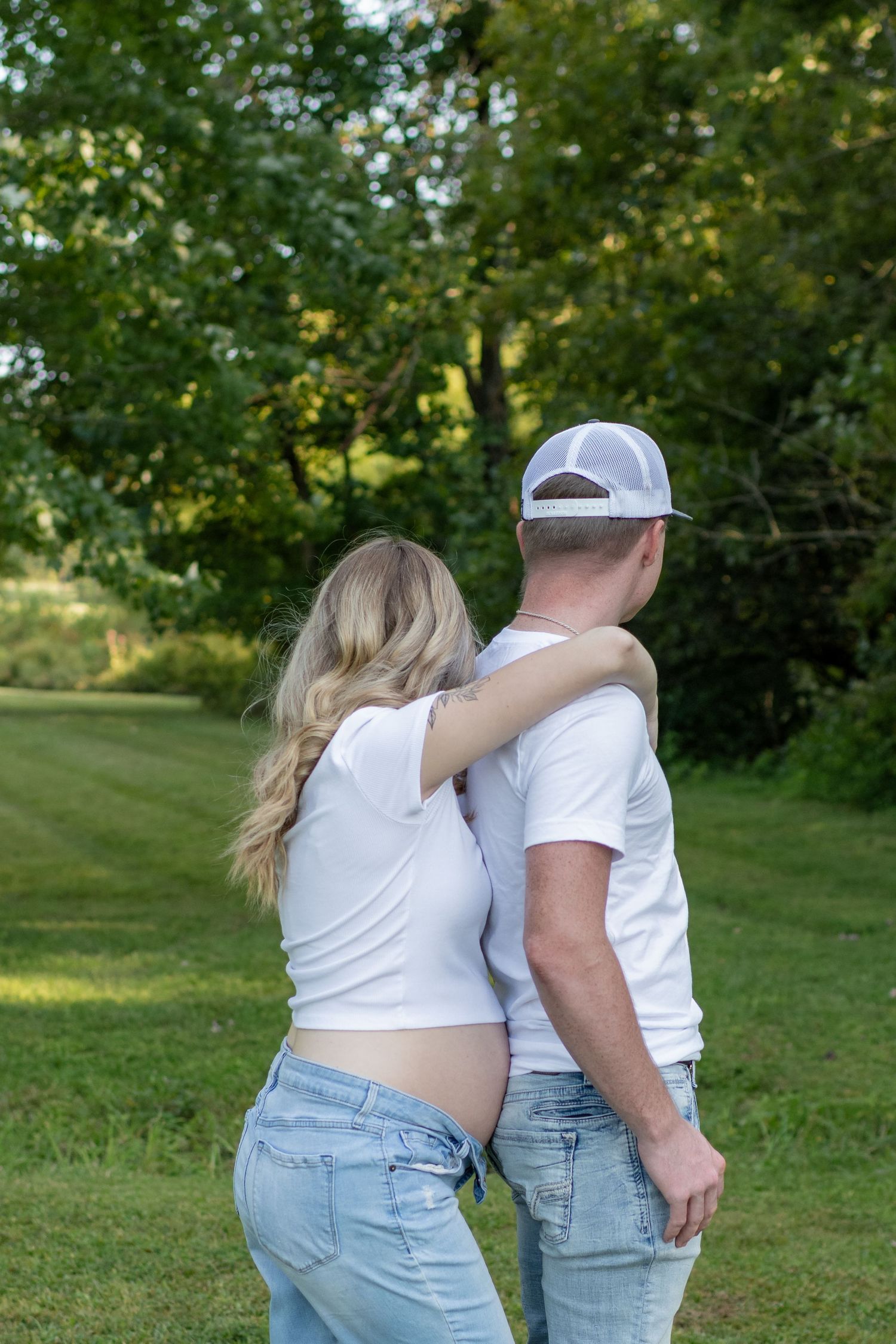 Pregnant couple in white shirts standing back-to-back viewing lush green trees.