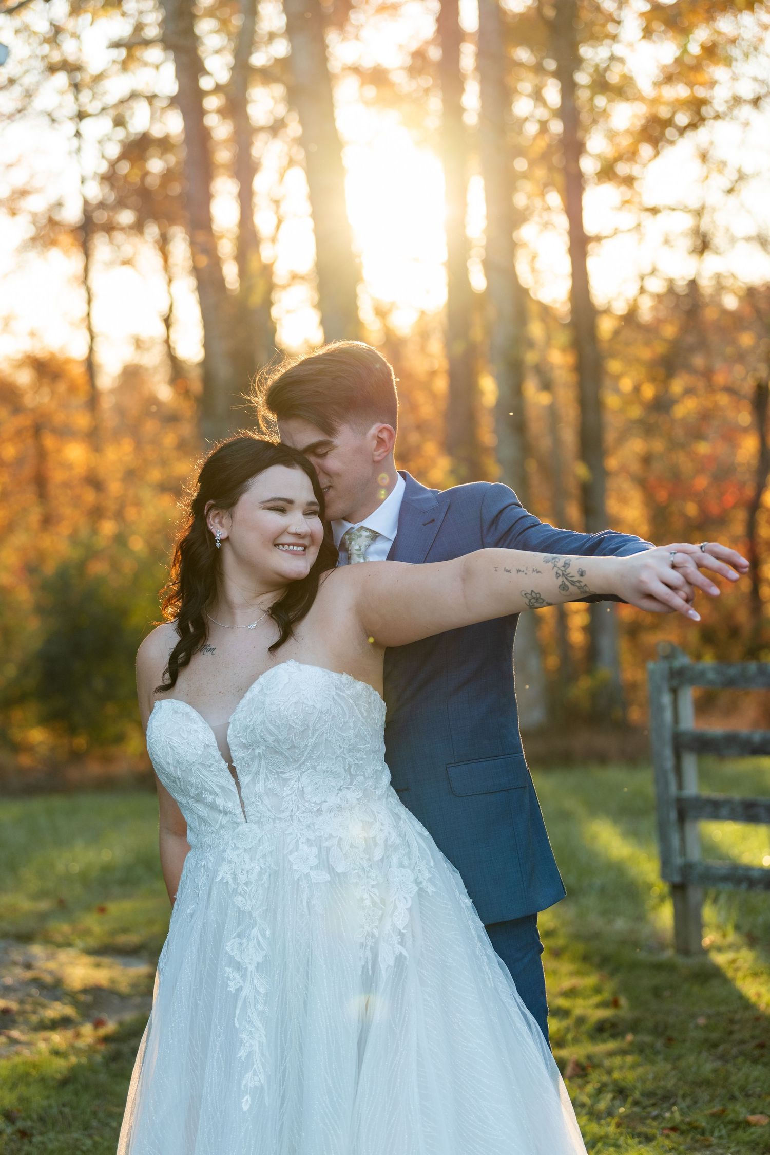 Bride and groom embrace at golden hour sunset in autumn forest, bride in lace ballgown, groom in navy suit.