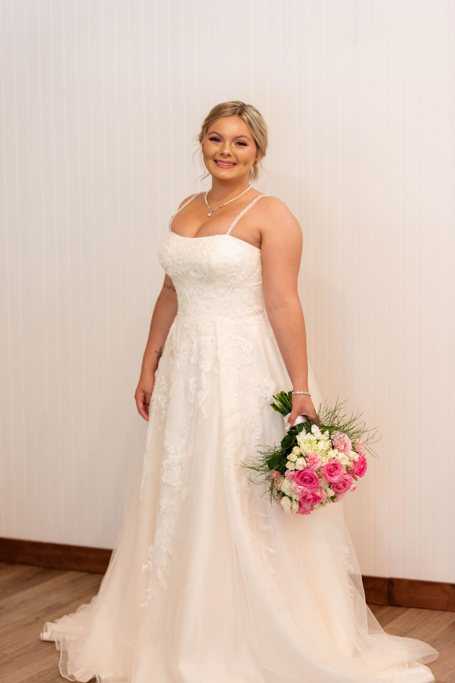 Elegant bride in lace spaghetti strap wedding gown holding pink rose bouquet indoors.