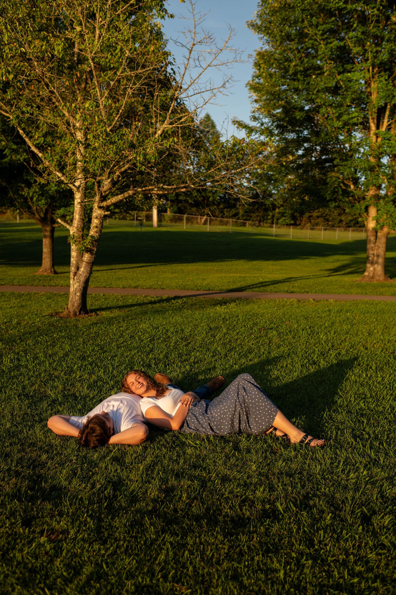 Couple lying together on lush green grass in a sunlit park, relaxing under golden hour light.