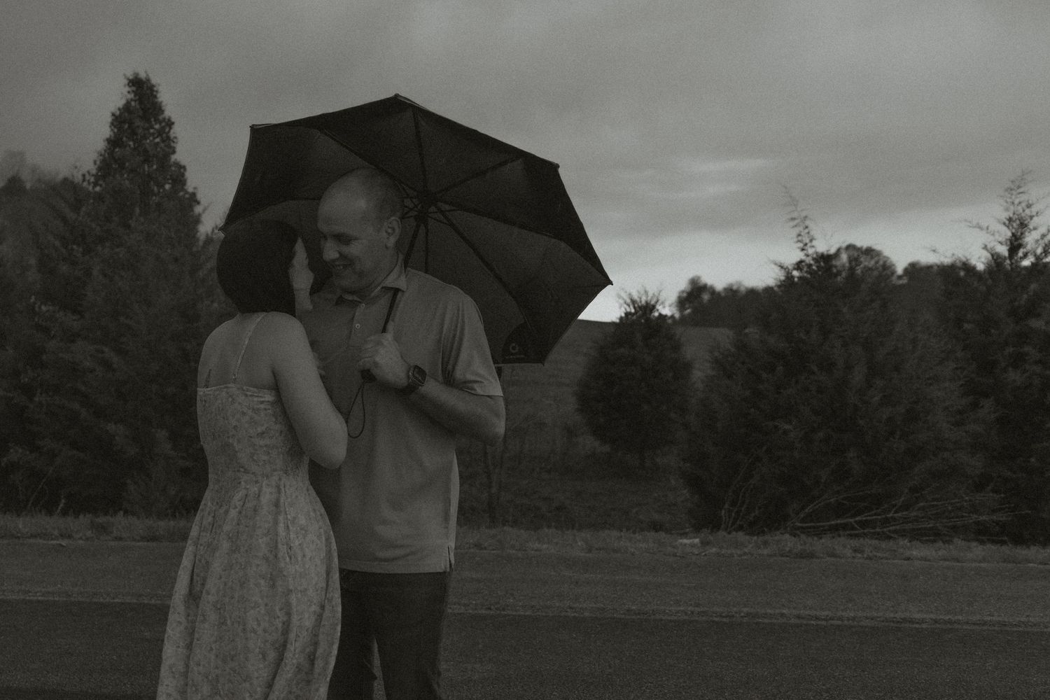 Black and white photo of a couple sharing an umbrella in a rainy outdoor setting.