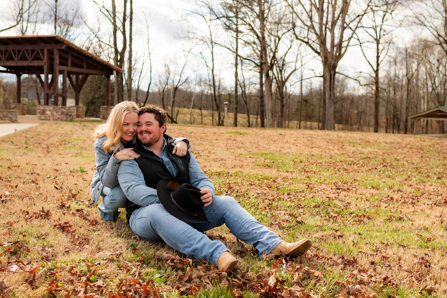 Happy couple sitting together on grass in a park, woman embracing man wearing a cowboy hat.