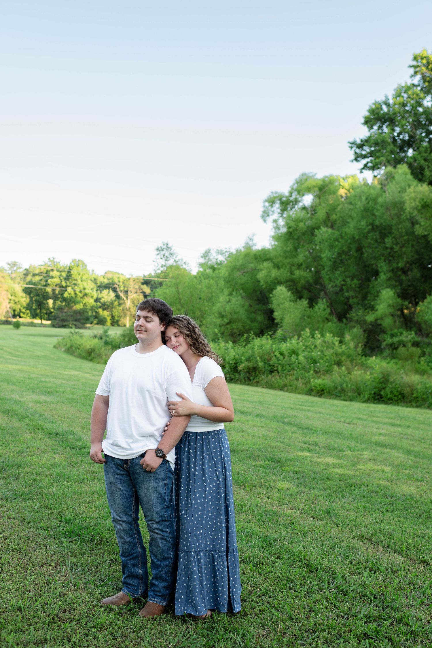 Couple standing together in green meadow, woman leaning head on mans shoulder affectionately.