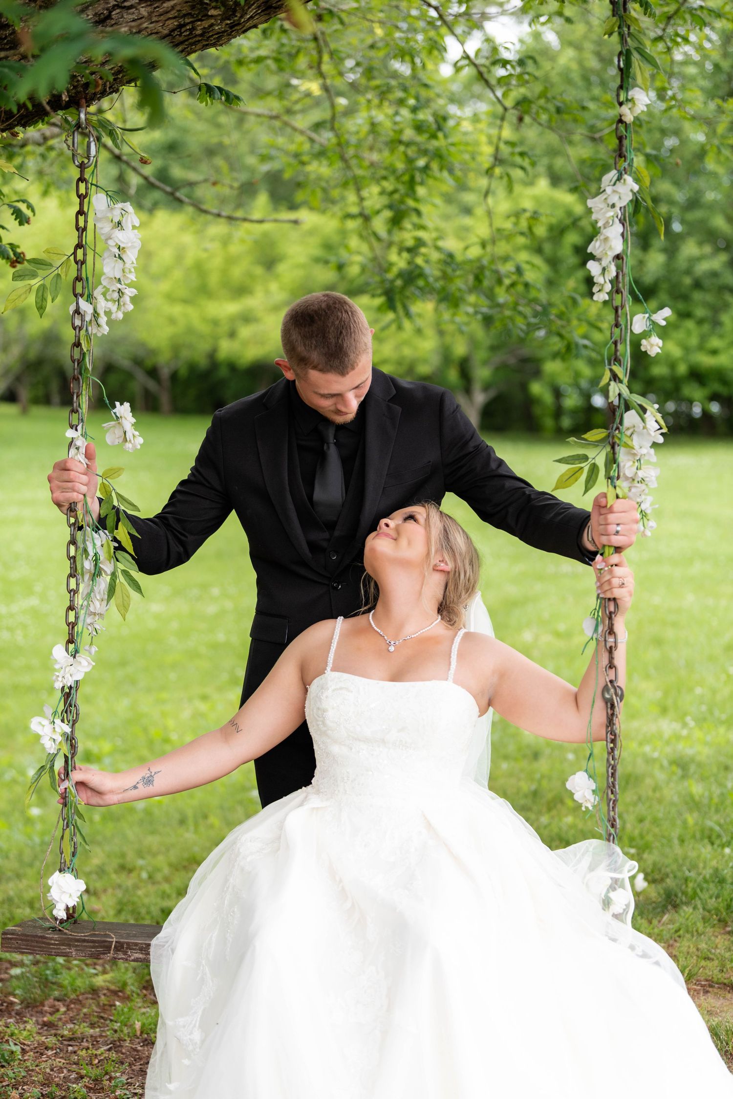 Groom stands behind bride seated on a flower-adorned swing, both laughing joyfully in an outdoor garden setting.
