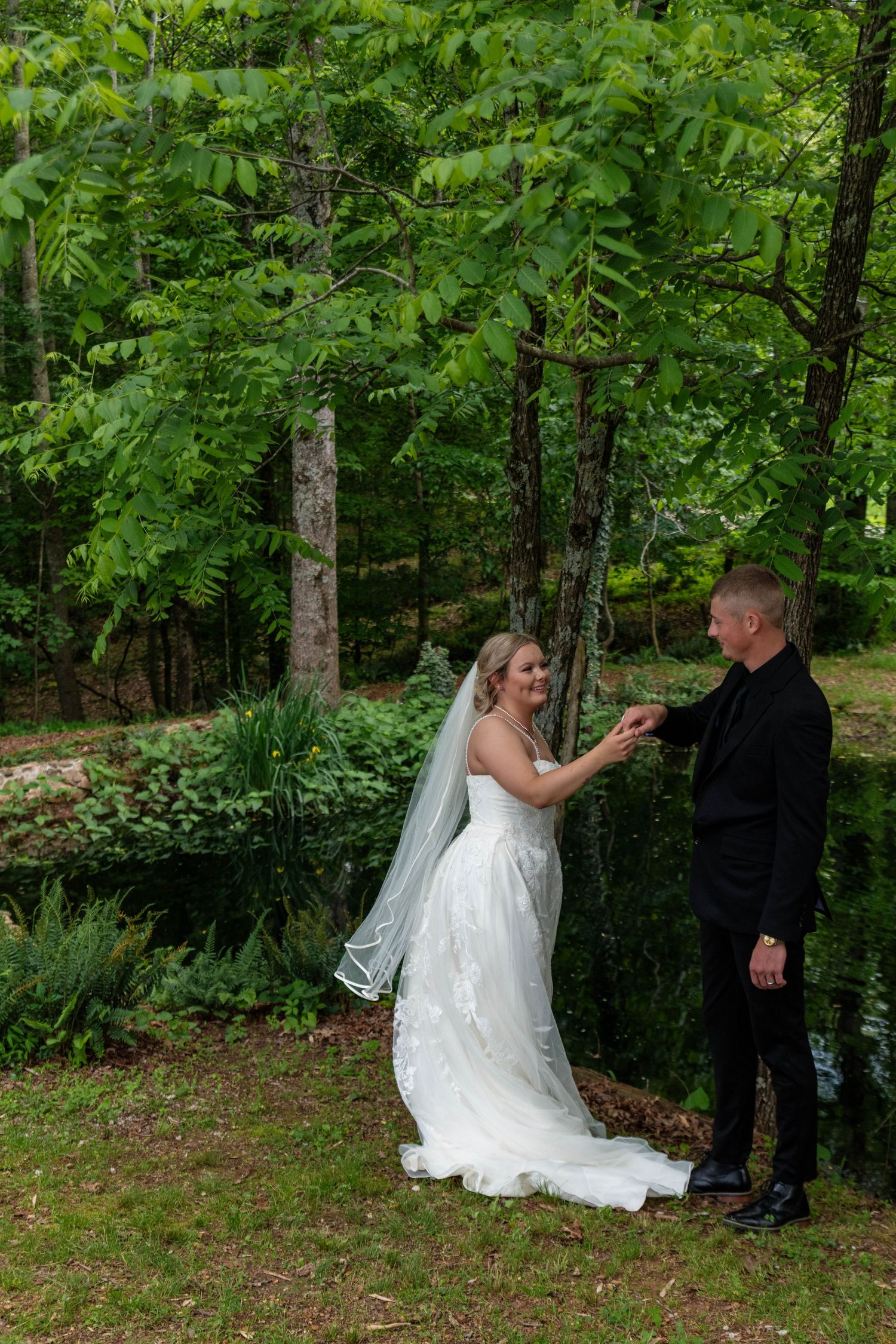Bride and groom holding hands near a lush green forest pond during outdoor wedding portraits.