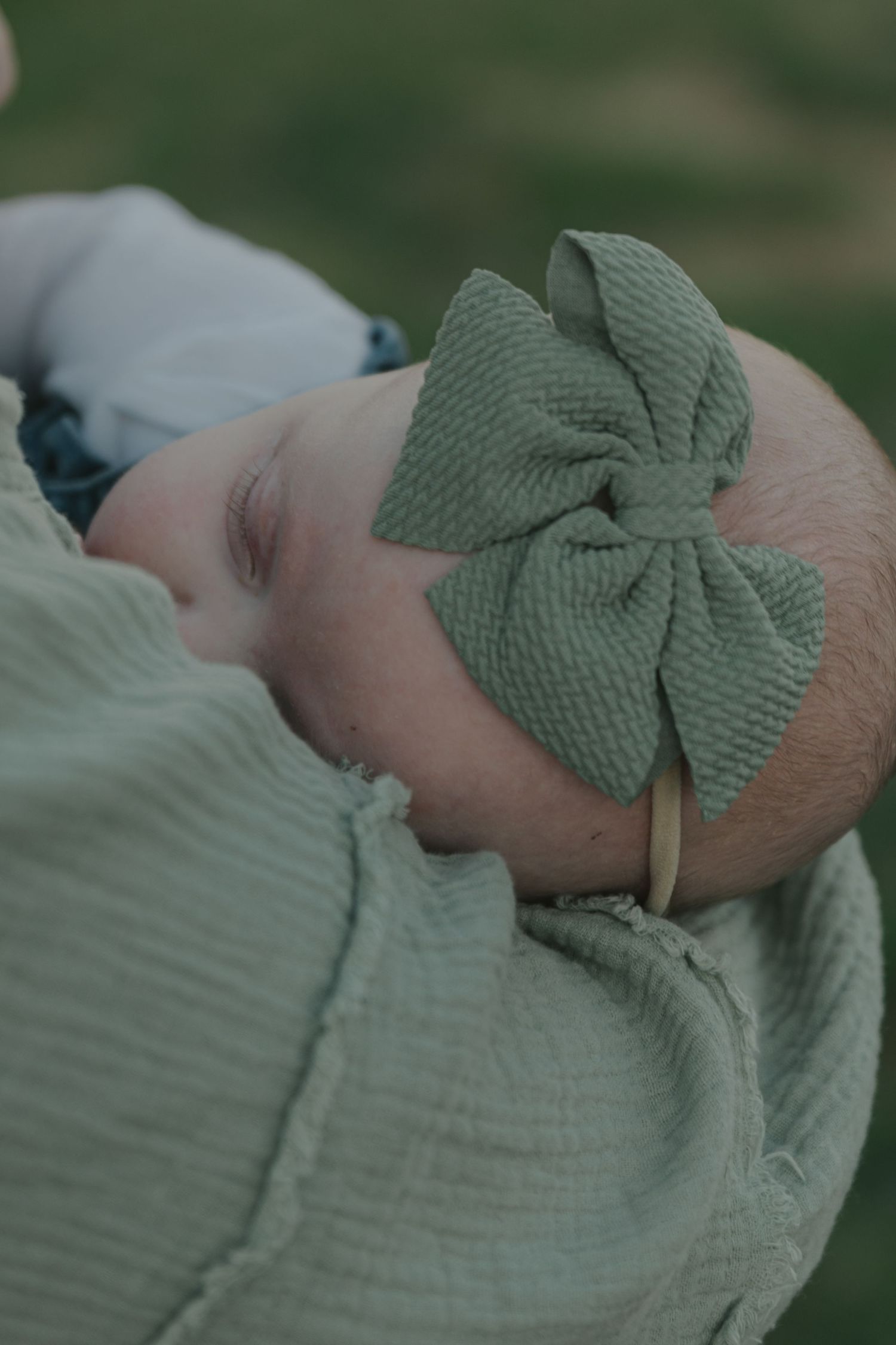 Close-up portrait of sleeping newborn baby wearing sage green bow headband outdoors.
