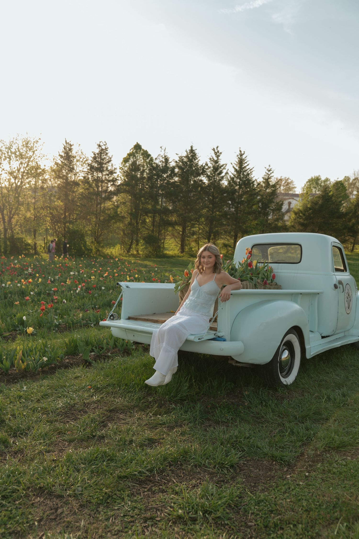 Blonde woman in white dress posing on vintage mint pickup truck in tulip field at sunset.