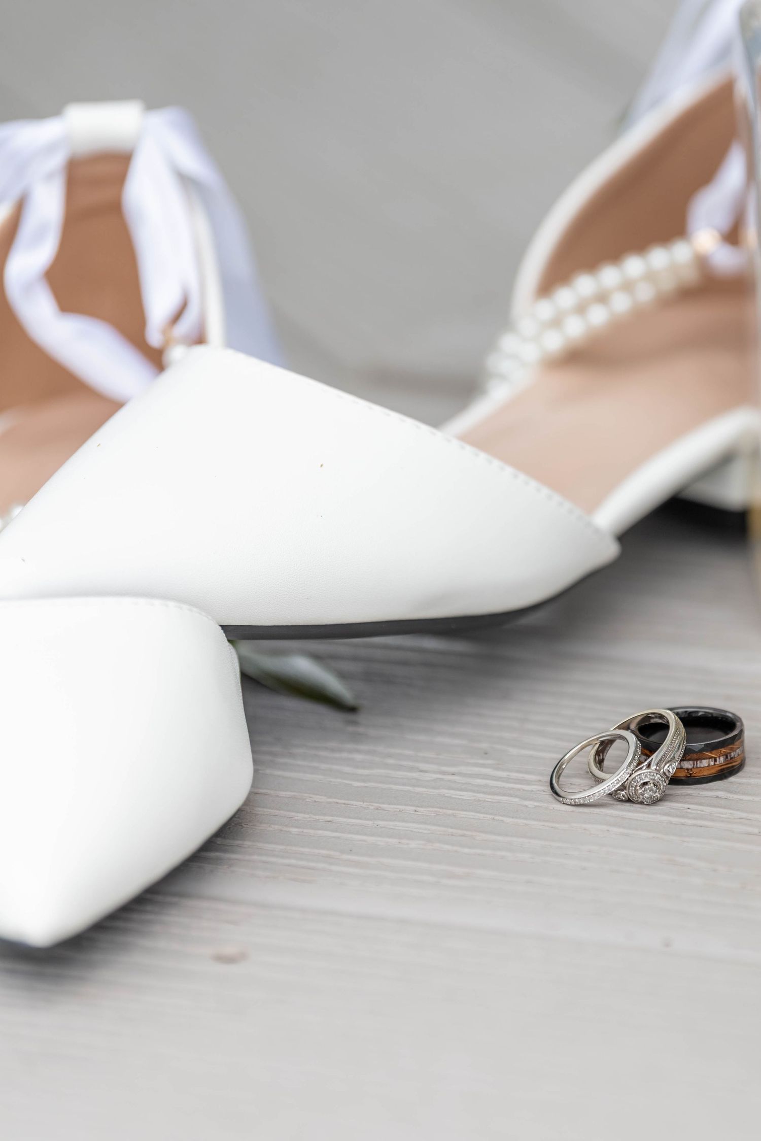 White pointed-toe bridal heels with pearl ankle straps beside wedding rings on a light wood surface.