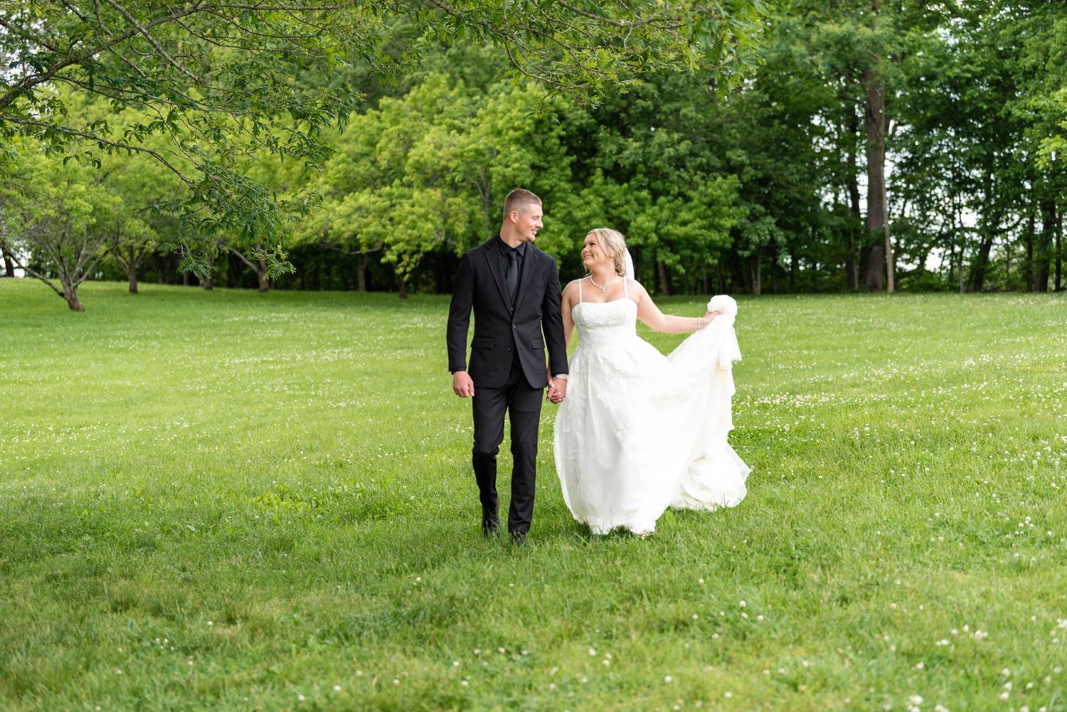 Bride and groom walk hand in hand across a lush green lawn, smiling at each other in wedding attire.