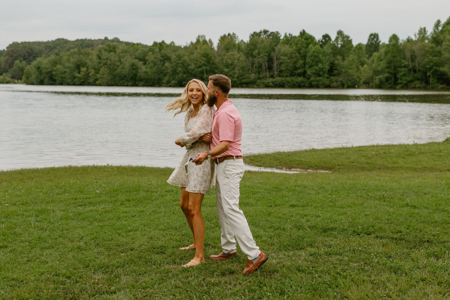 Happy couple walks hand in hand along a green lakeside lawn with trees in the background.