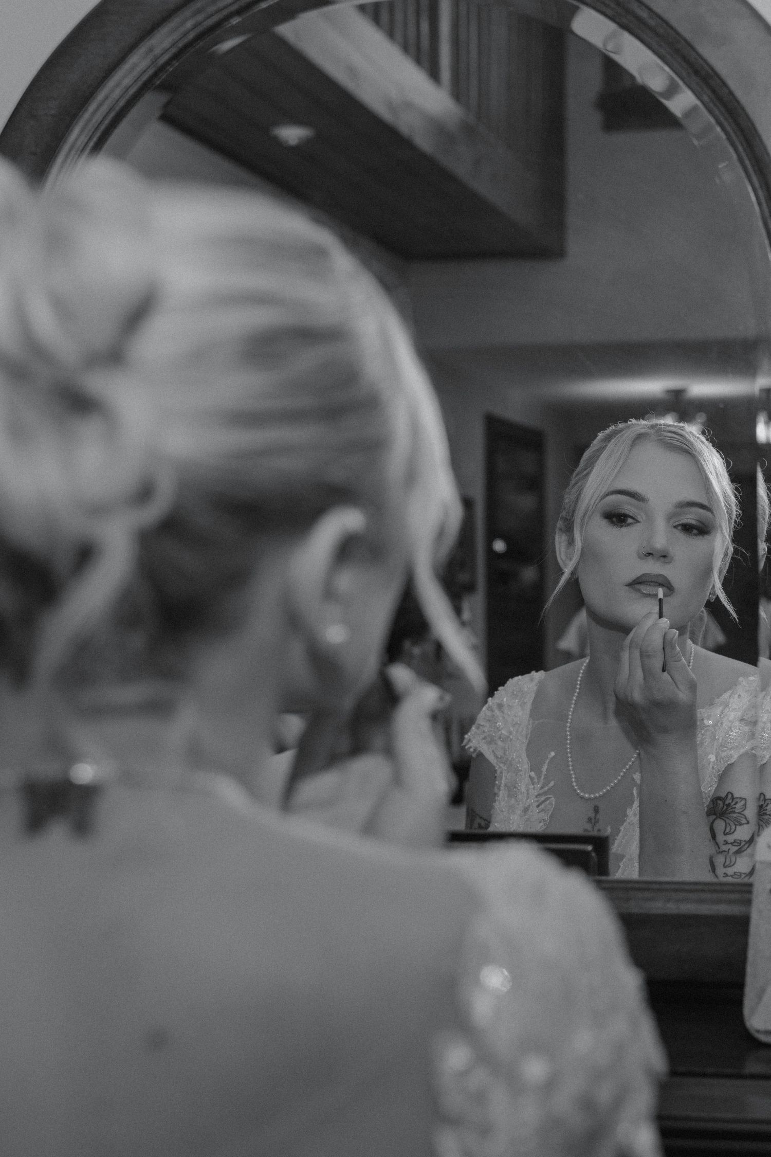 Black and white photo of bride applying lipstick while looking into round mirror before wedding.