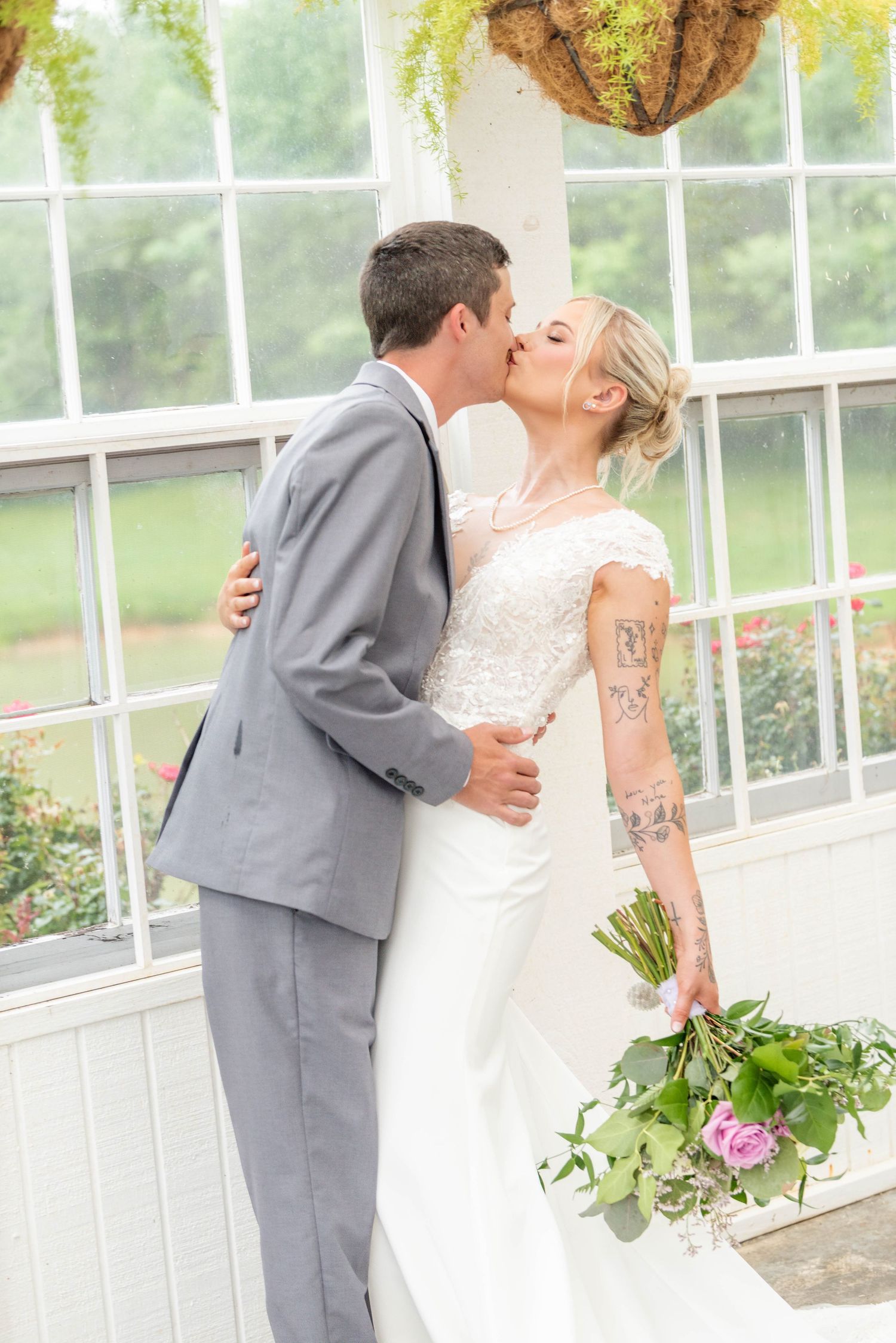 Bride and groom share a passionate kiss in a bright greenhouse as bride holds a floral bouquet.