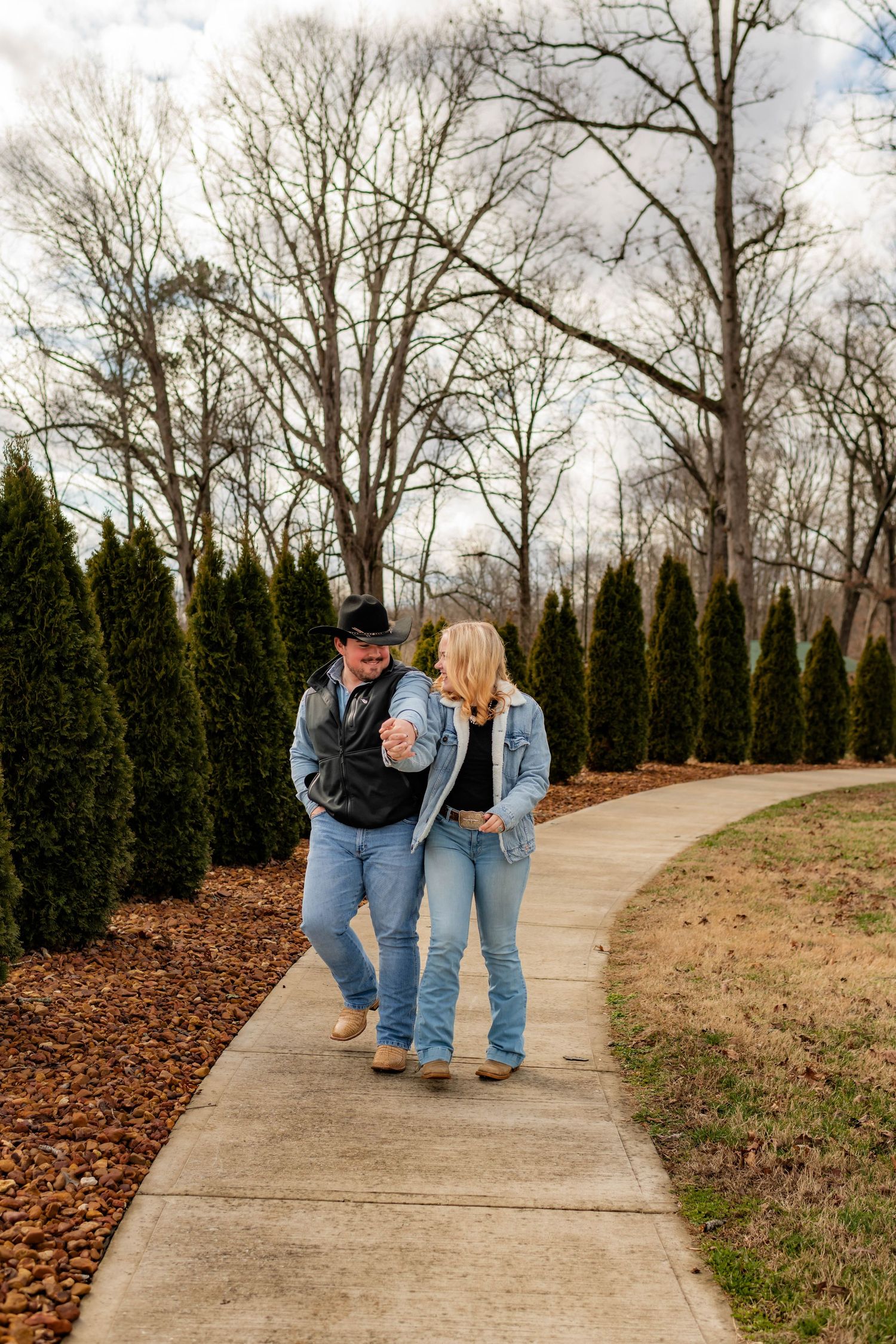 Couple walking hand-in-hand on a curved sidewalk lined with evergreen trees in a park.