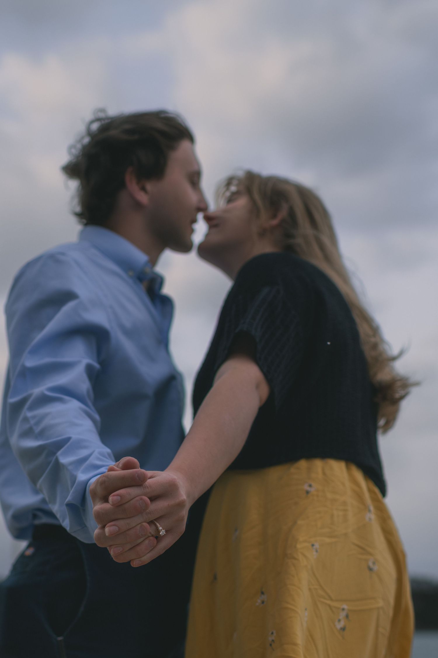 Romantic couple leaning in for a kiss while holding hands under a cloudy sky.