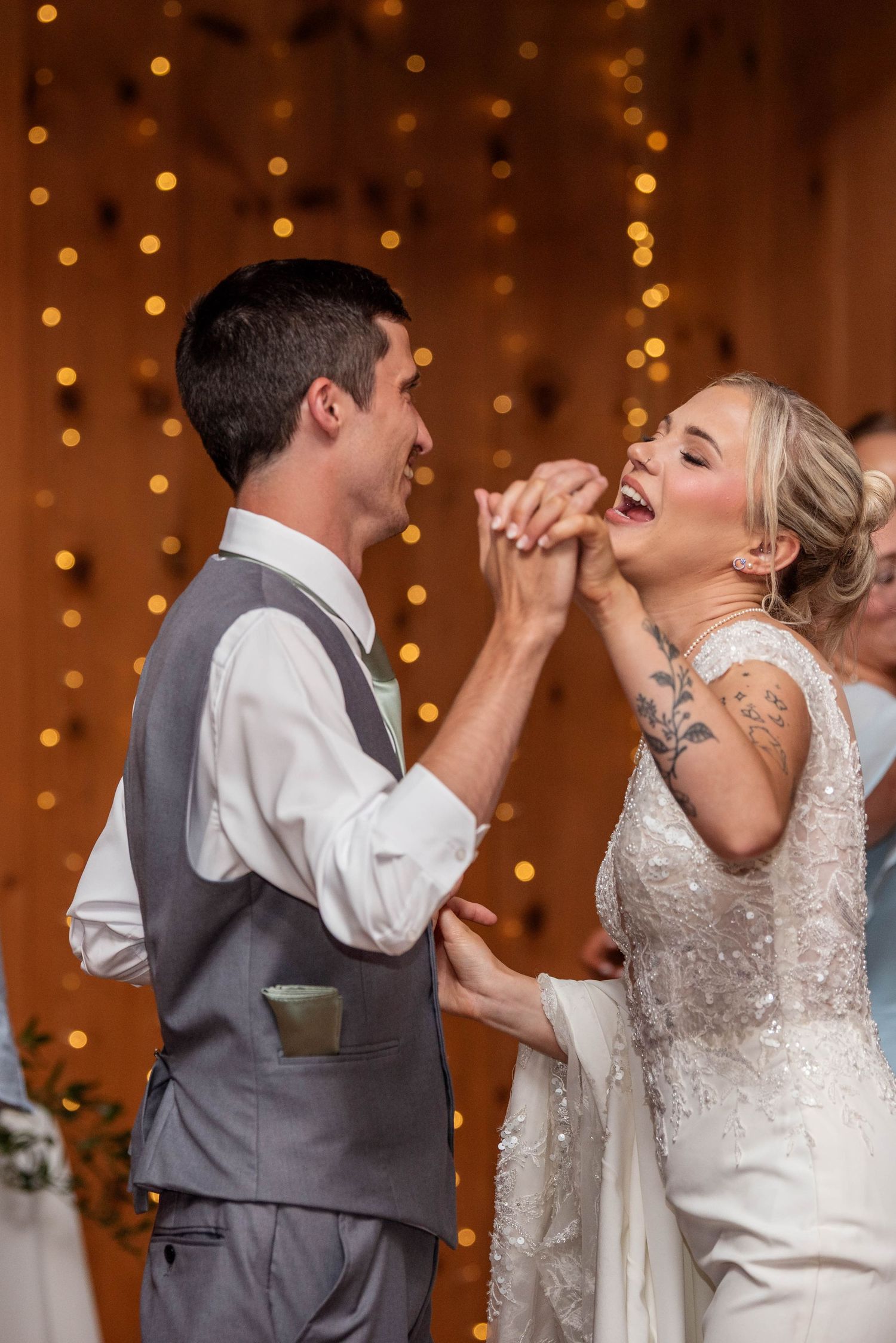 Joyful bride and groom laugh together while feeding cake during their wedding reception with bokeh lights.