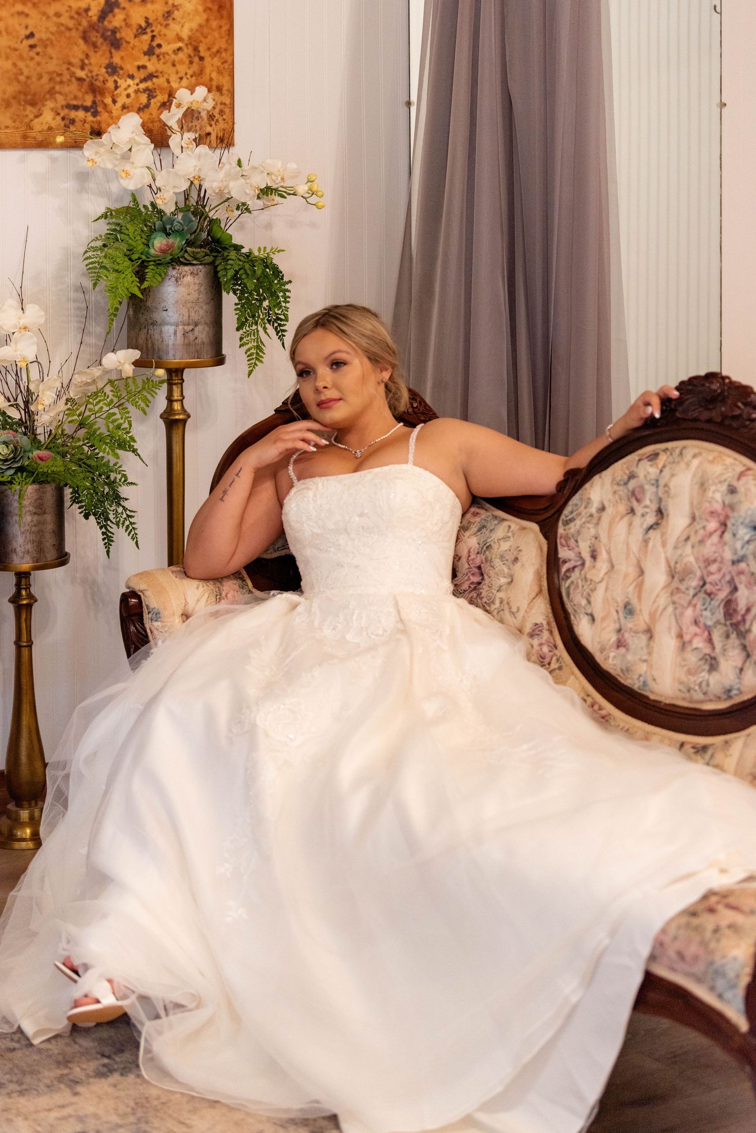 Elegant bride in a white lace gown poses gracefully on an ornate vintage sofa in a beautifully decorated room.