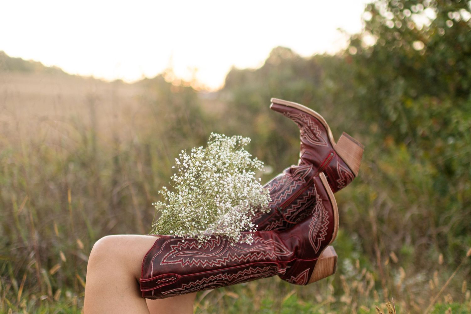 Legs raised in decorative brown cowboy boots with baby's breath flowers tucked inside, set against a golden field at sunset.