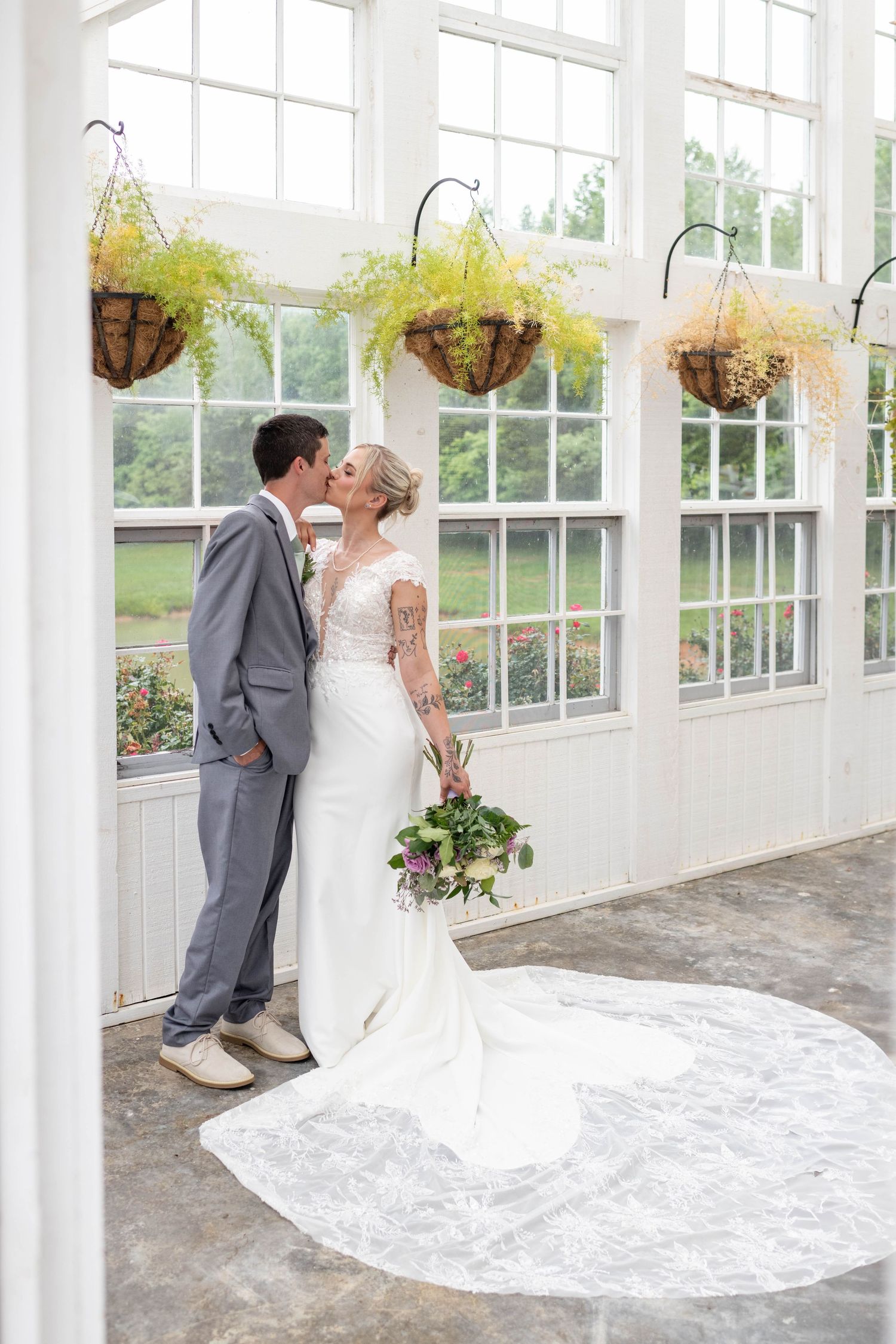 Bride and groom share a kiss inside a bright greenhouse with hanging fern baskets and large windows.