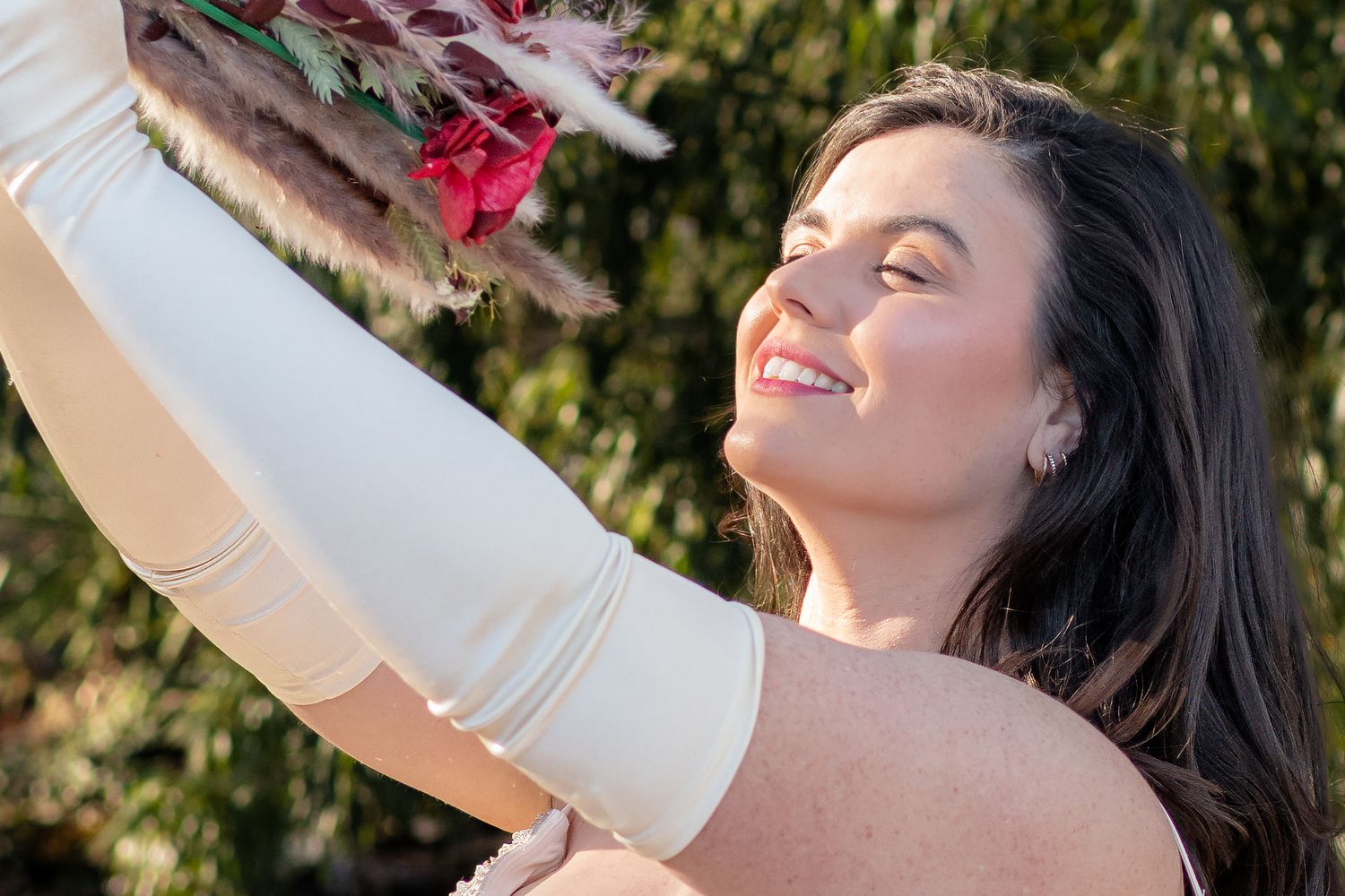 Smiling bride in white gown and gloves holds a floral bouquet outdoors in sunlight.