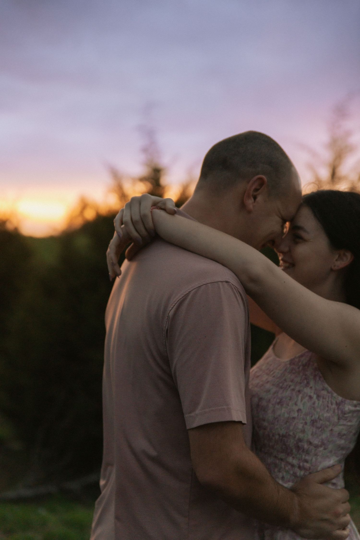 Couple tenderly touching foreheads and embracing against a beautiful purple and orange sunset sky.