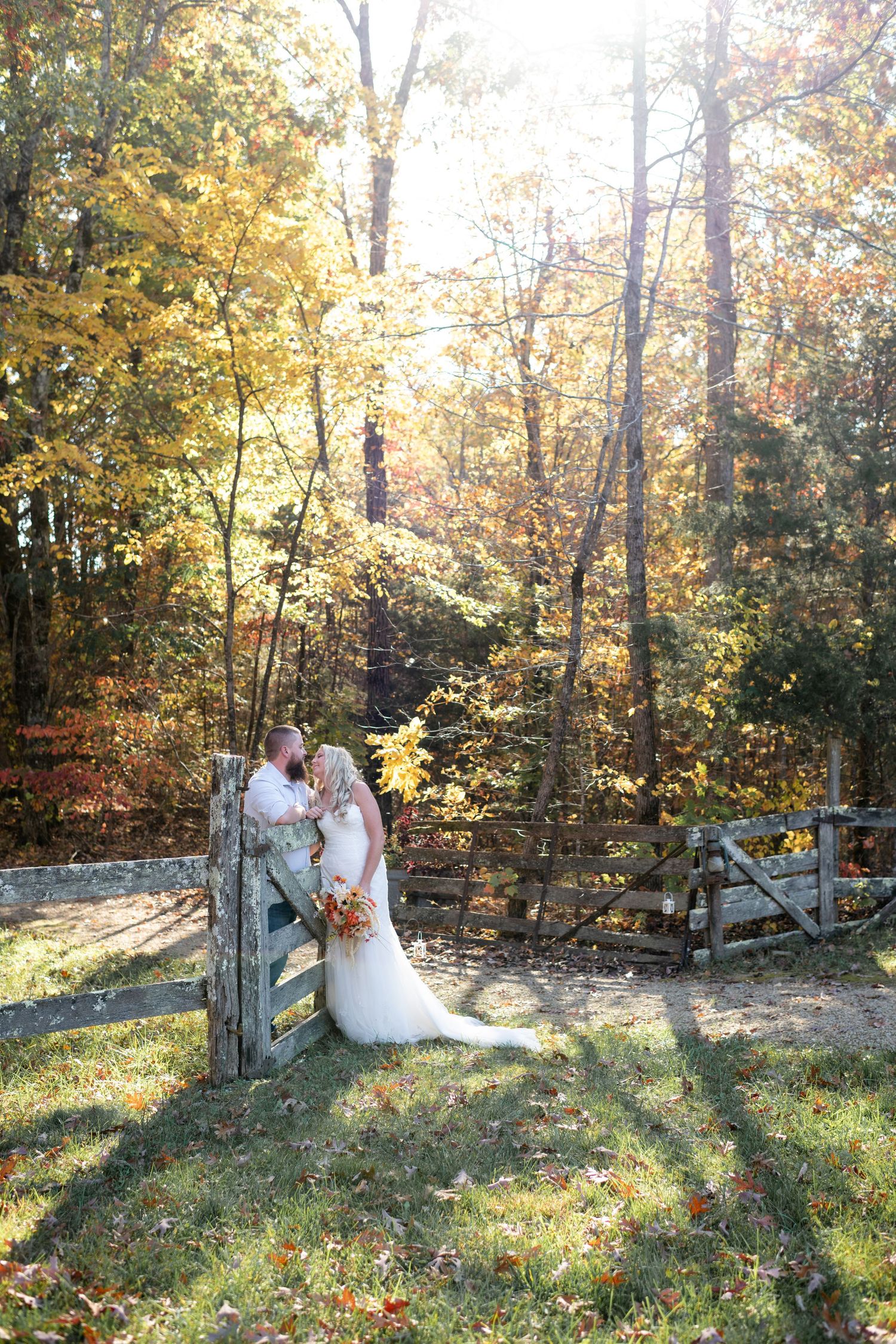 Bride and groom embrace near a rustic wooden fence surrounded by golden autumn forest sunlight.