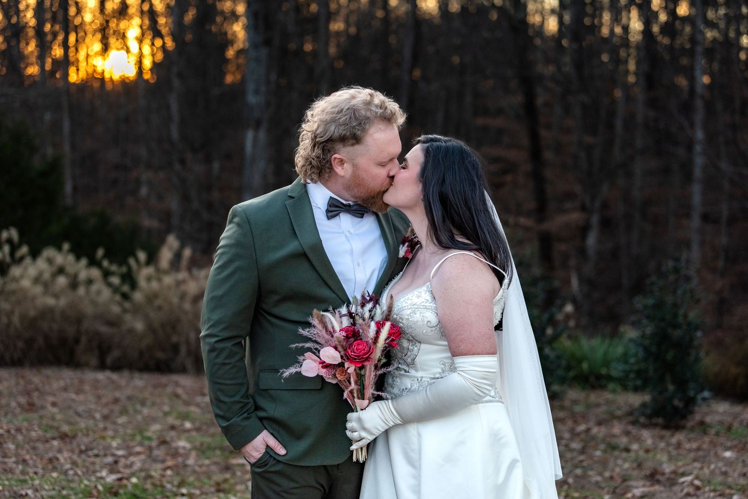 Newlywed couple kissing at sunset with bride holding red bouquet in a forest setting.