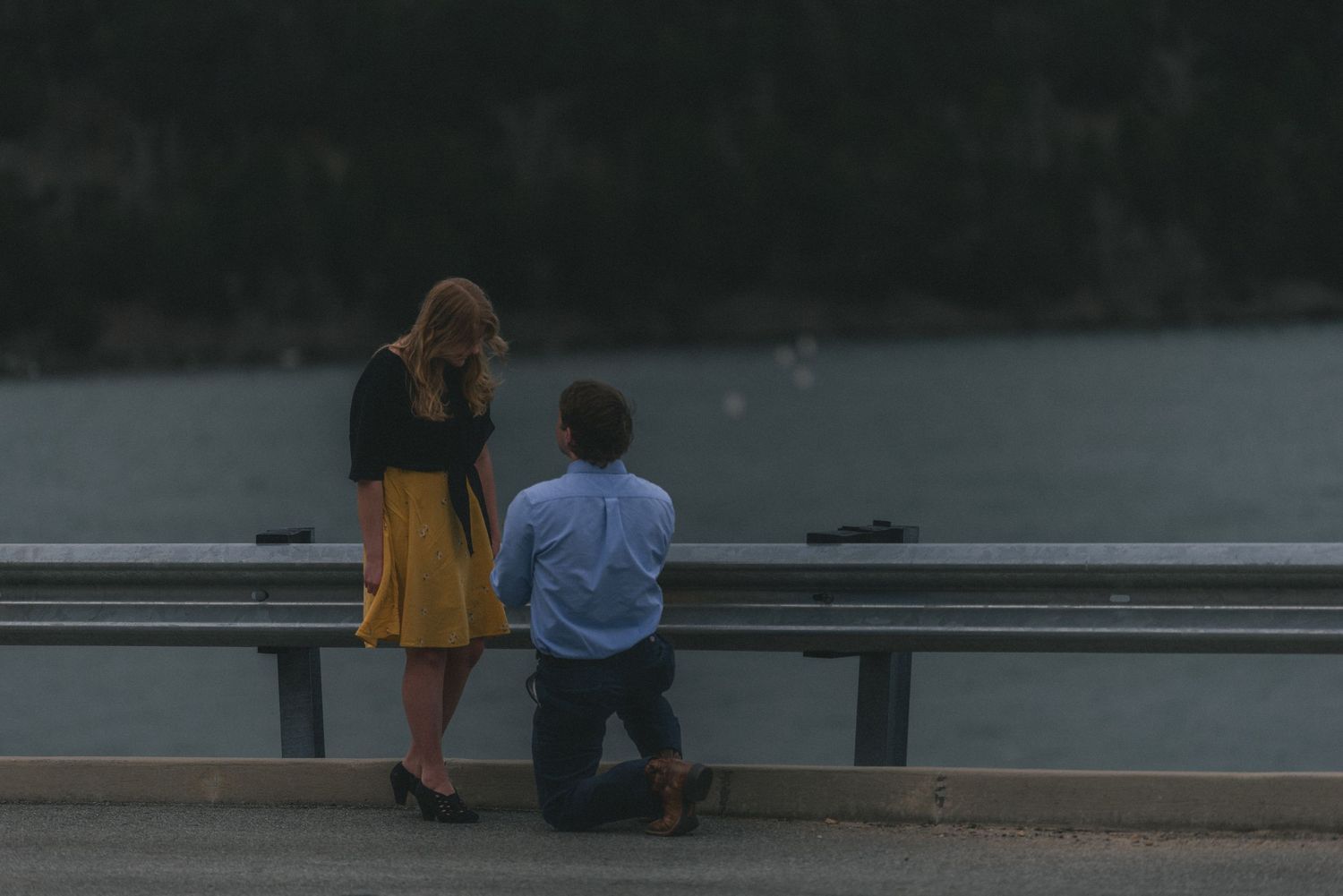 Man proposing on one knee to woman by a waterside guardrail at dusk.