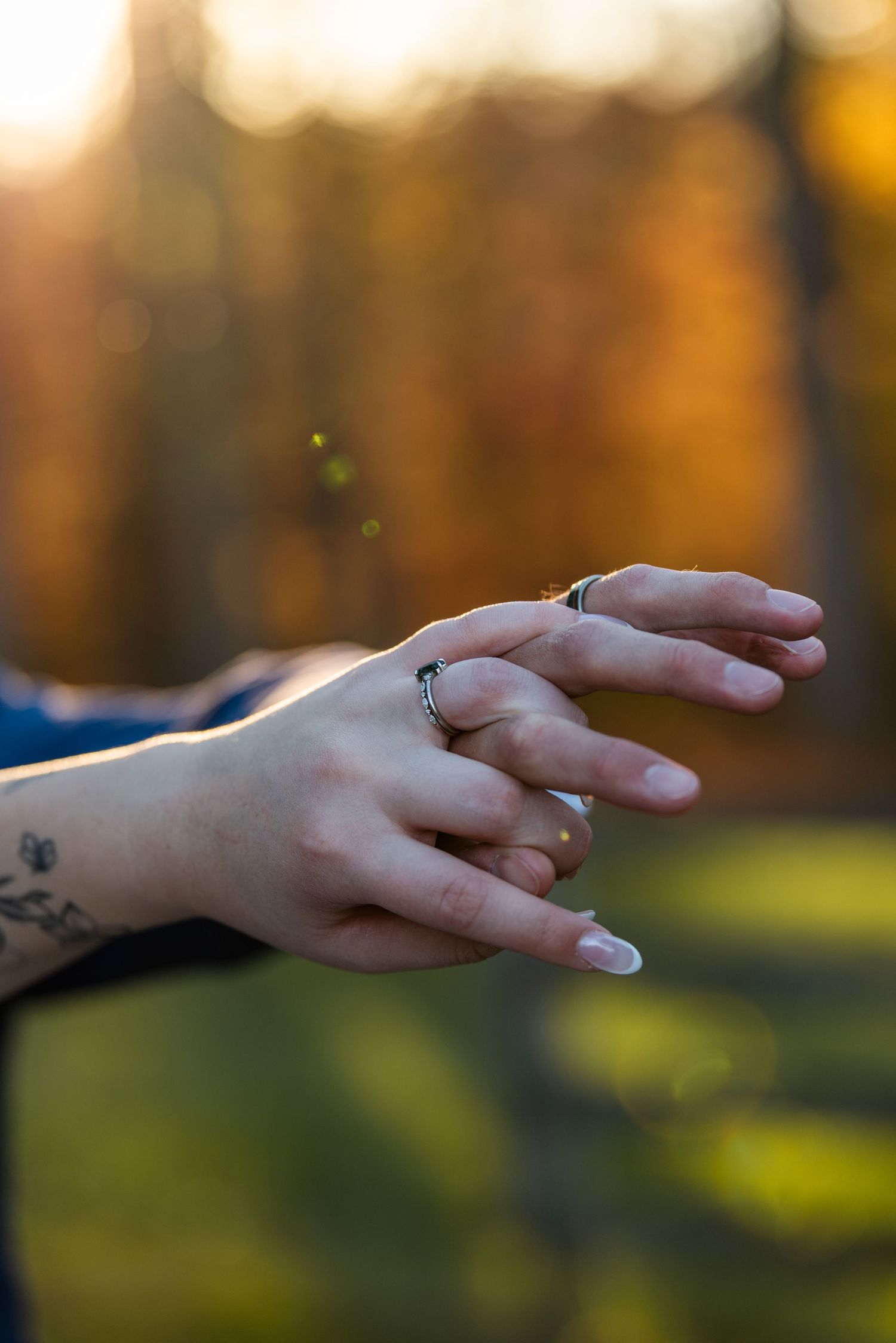 Close-up of bride and groom hands with wedding rings and tattoo at golden hour outdoor ceremony.