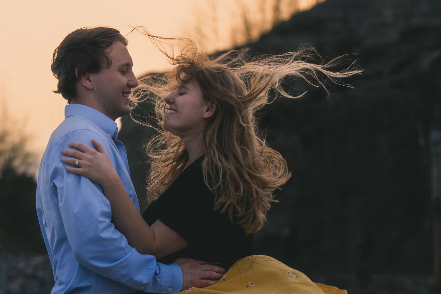 Couple embracing at sunset with wind blowing woman's hair, romantic outdoor engagement photo.