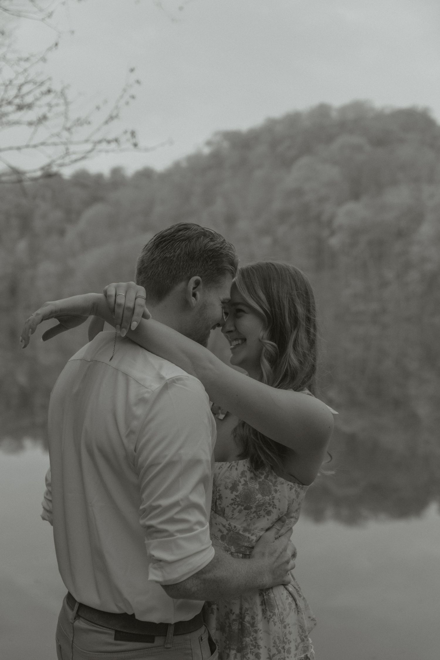 Black and white photo of a couple embracing and smiling at each other near a reflective lake and wooded hillside.