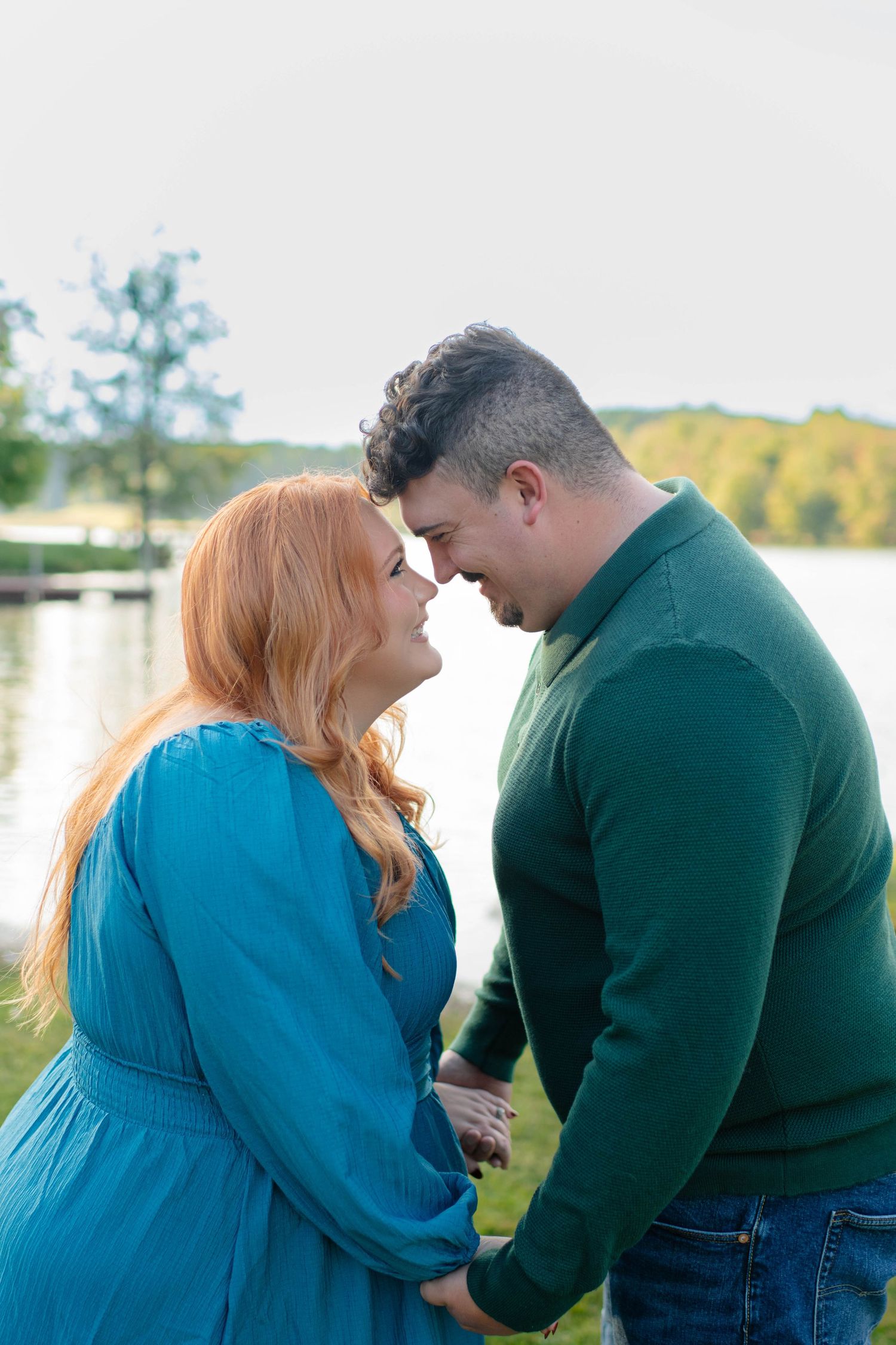 Romantic couple touching foreheads and holding hands by a serene lake with autumn trees in the background.