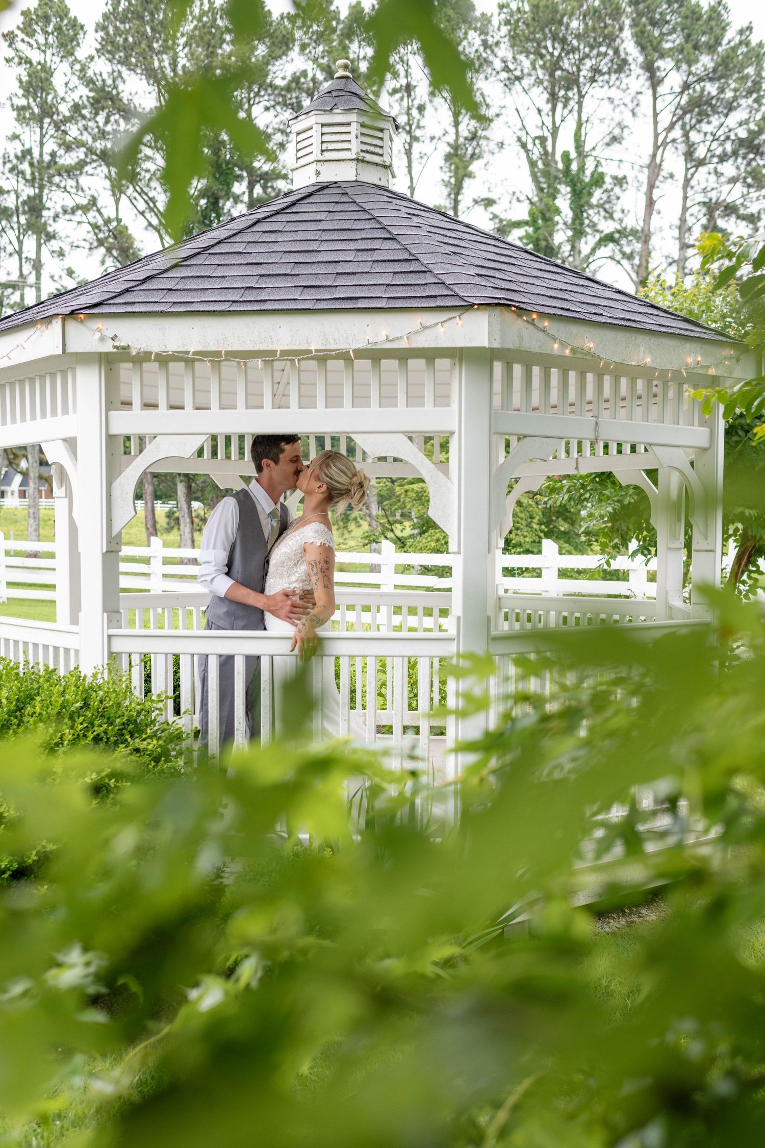Bride and groom kissing under white gazebo surrounded by lush green garden on wedding day.