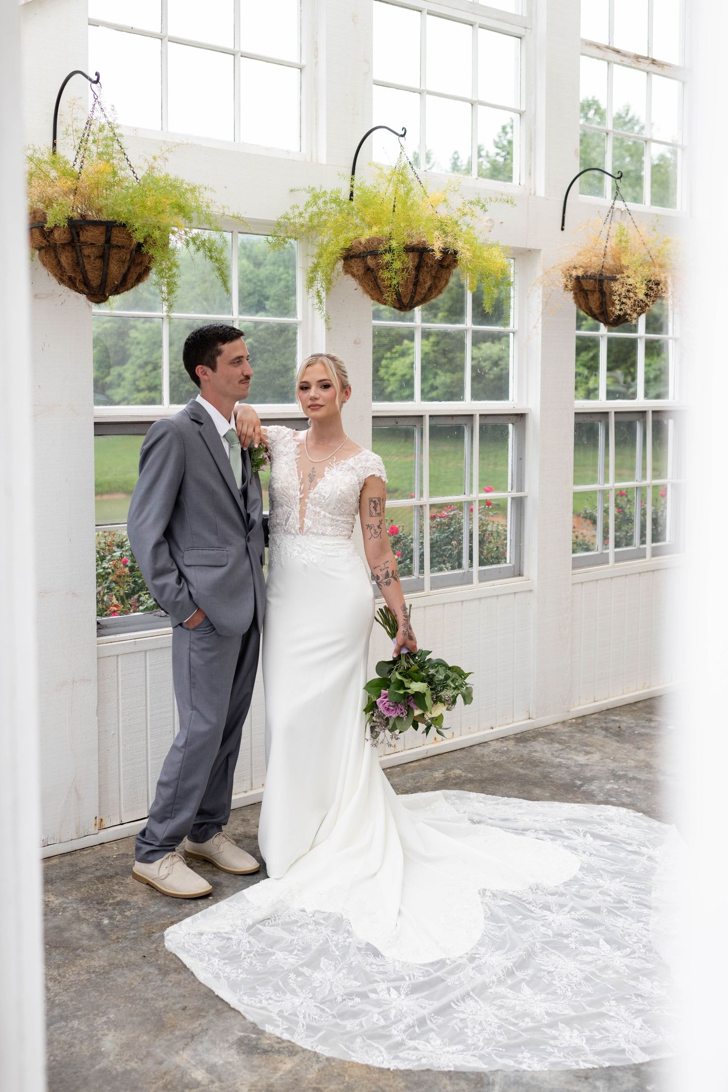 Bride and groom pose elegantly in a sunlit greenhouse with hanging ferns and large white-framed windows.