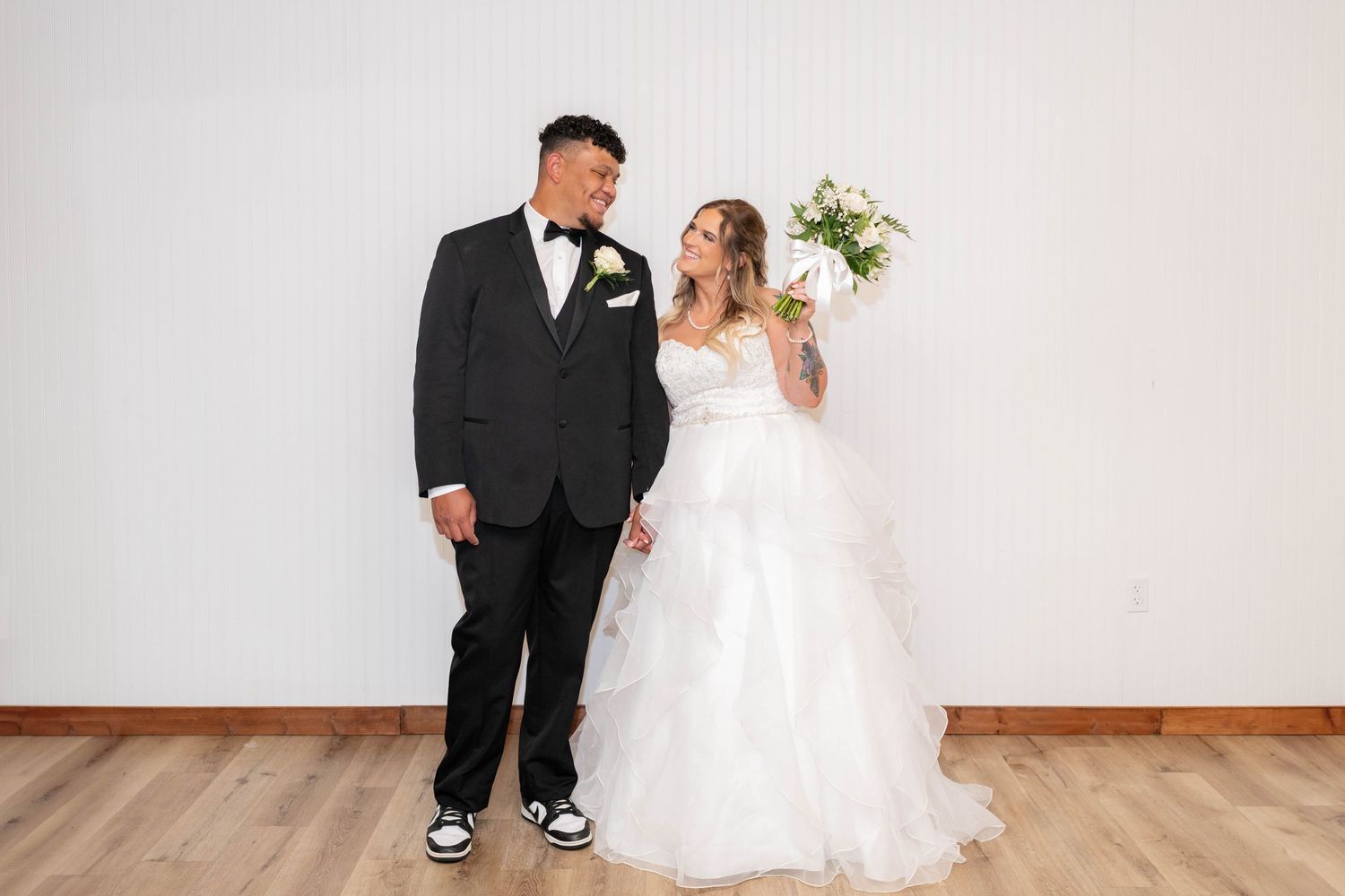 Bride and groom pose together, she holds a bouquet while he wears a black tuxedo with sneakers.