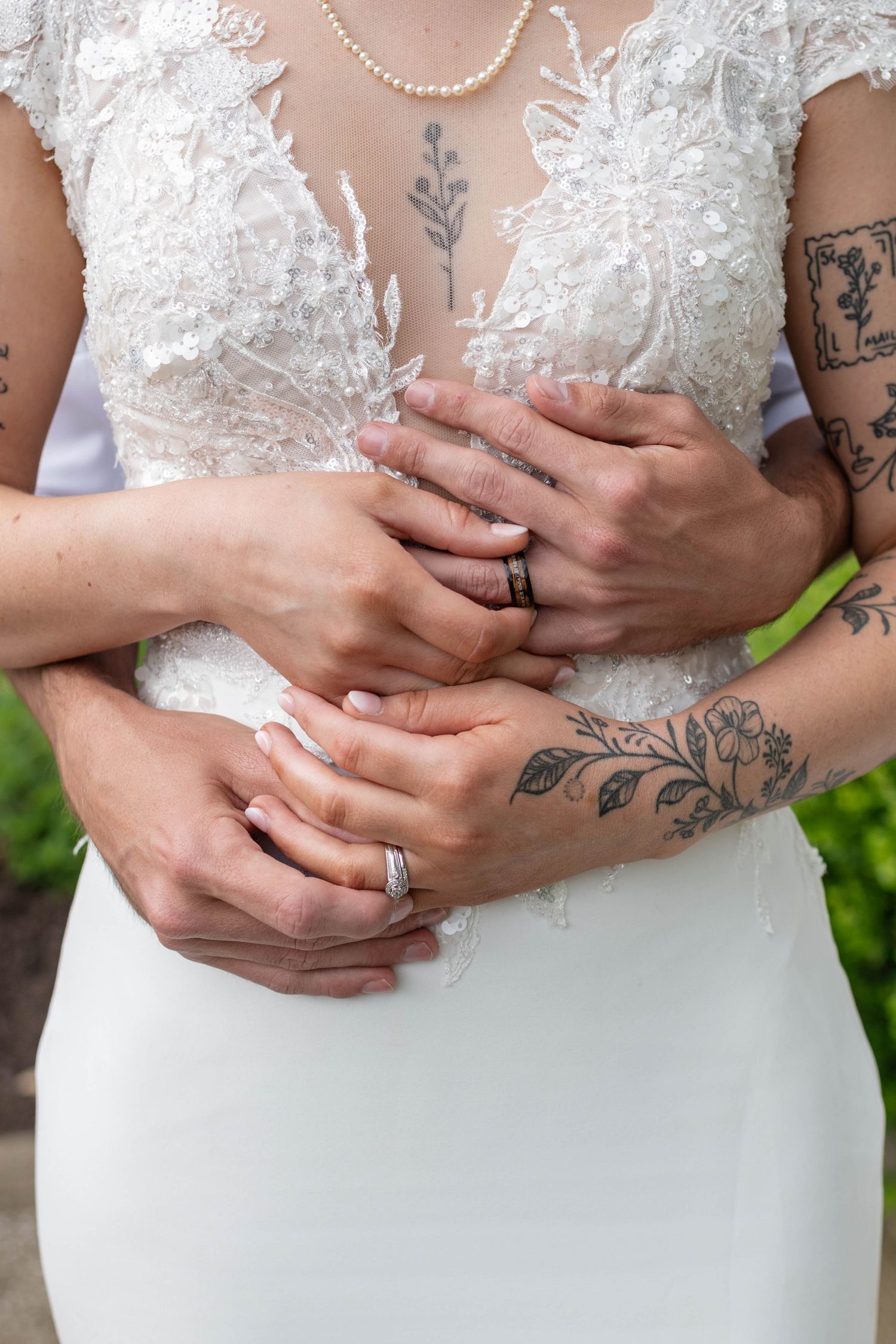 Close-up of bride and groom hands with wedding rings, bride in lace gown with floral tattoos.