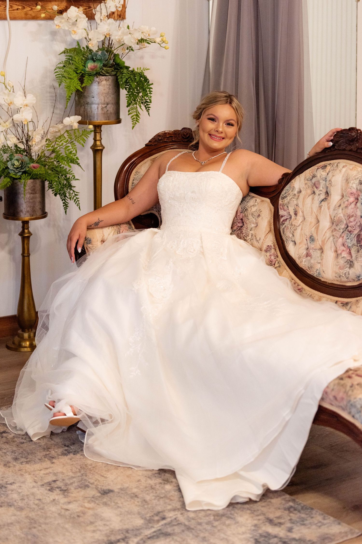 Bride in a strapless lace ball gown sitting elegantly on a vintage chair in a studio.