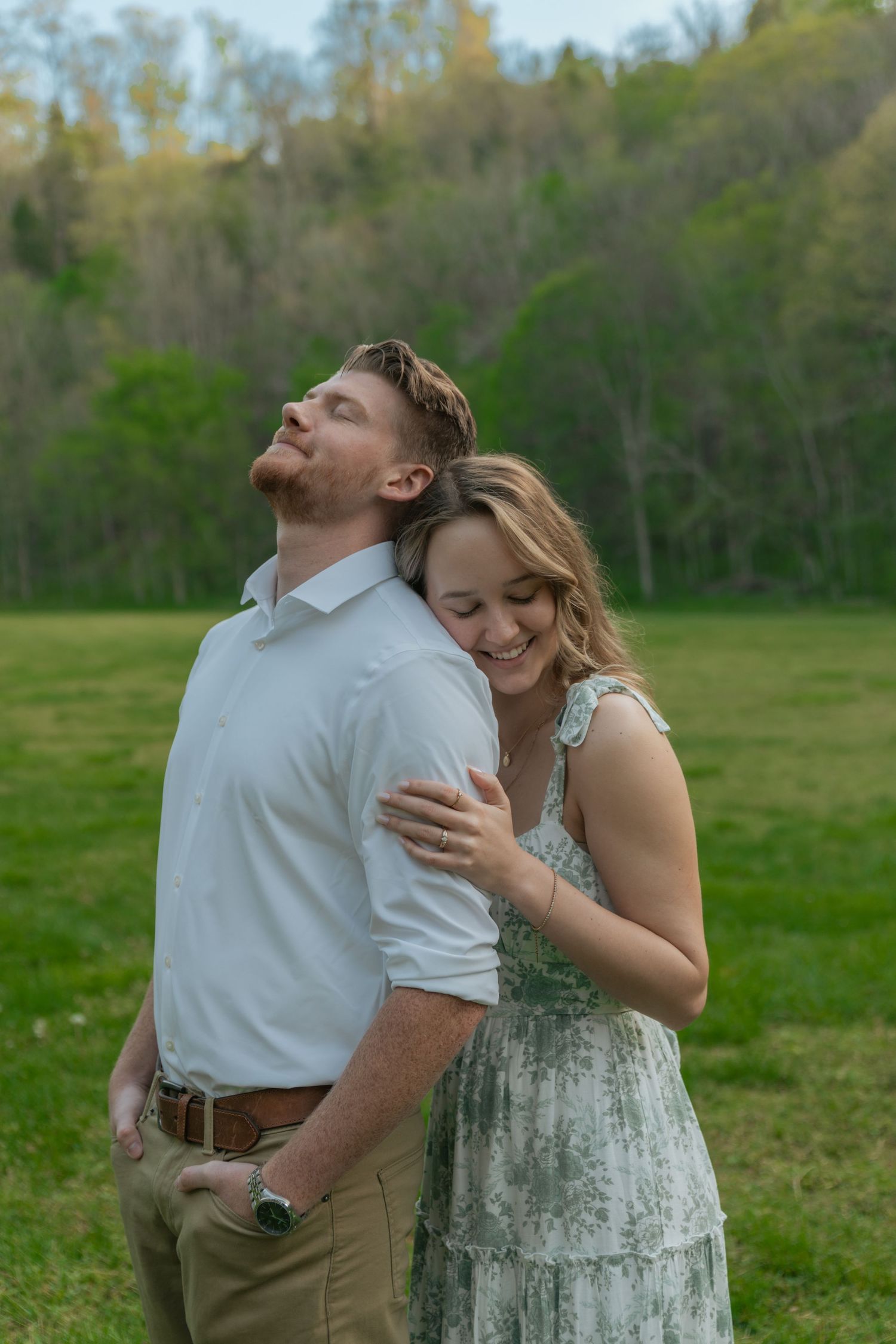 Joyful couple laughing together in a green meadow, woman hugging man from behind near spring forest.