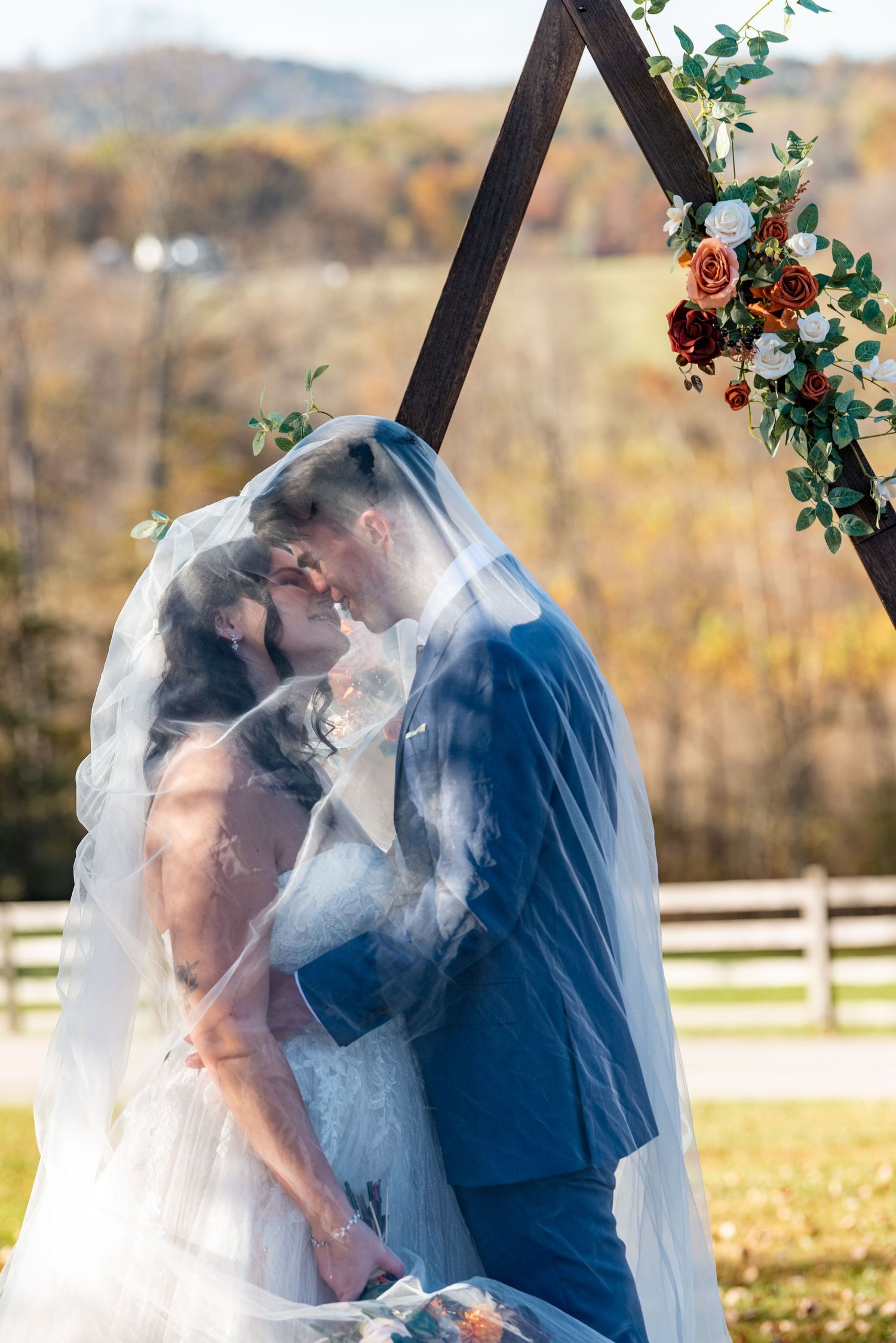 Bride and groom kissing under veil at outdoor fall wedding ceremony with floral arch.