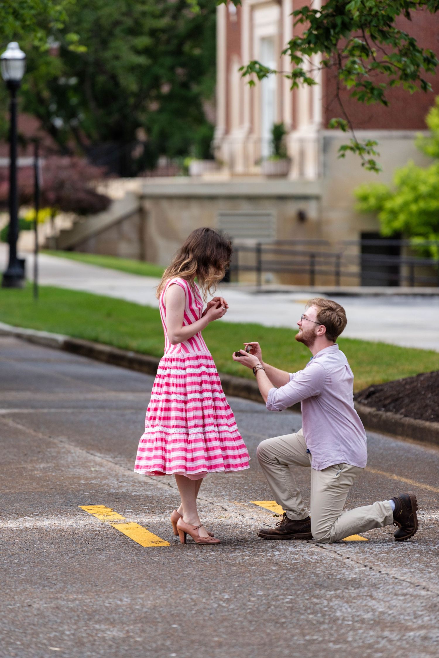 Man proposing on one knee to woman in pink dress on a tree-lined street during a surprise proposal.