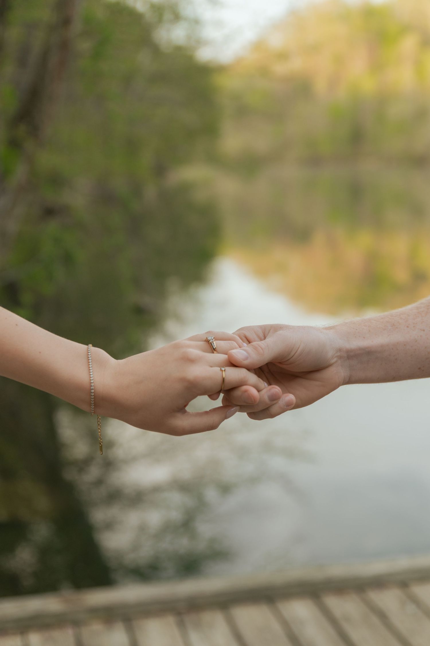 Close-up of engaged couple holding hands near a reflective pond, showing delicate rings and bracelet.
