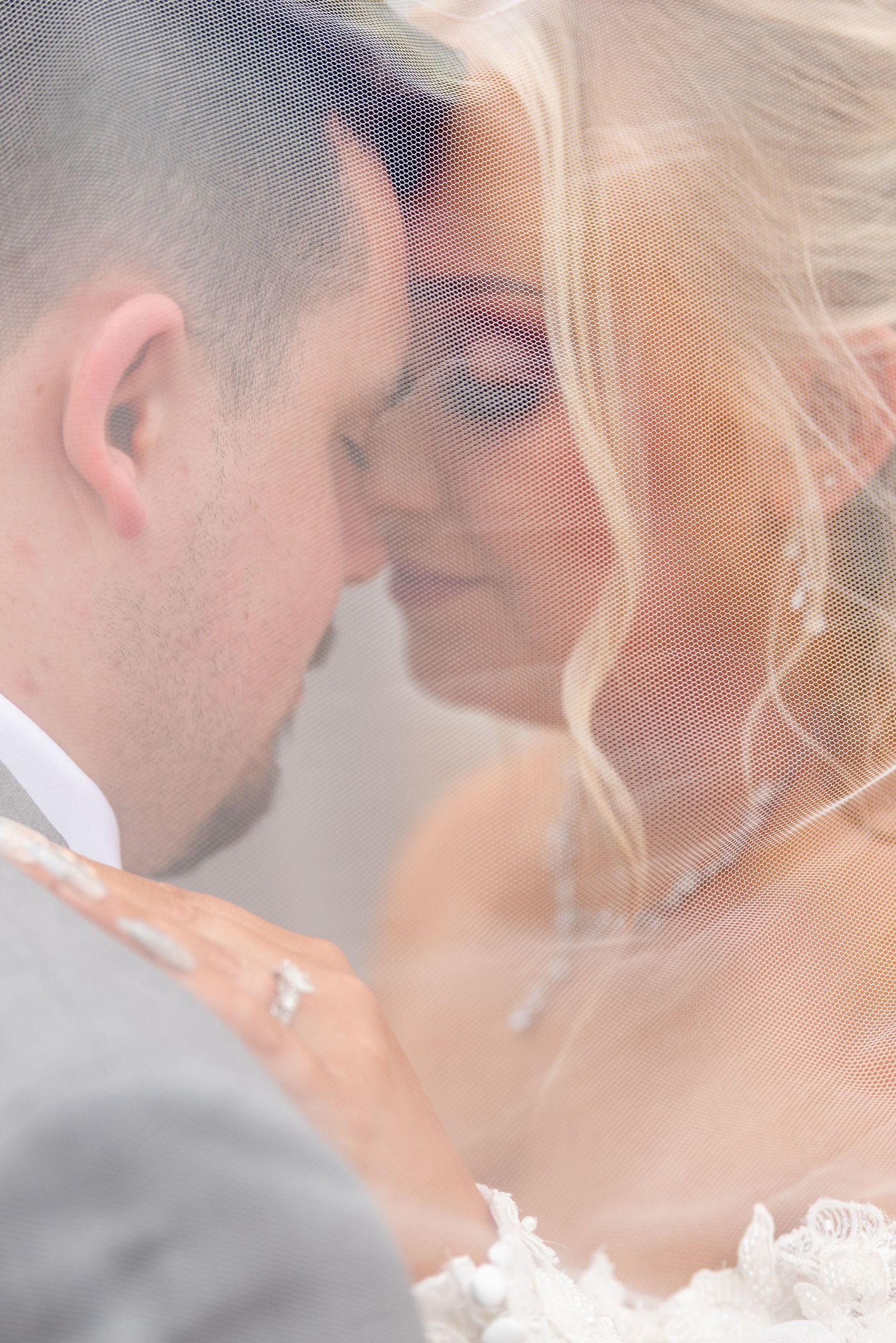 Bride and groom share an intimate moment under a flowing wedding veil, foreheads touching softly.