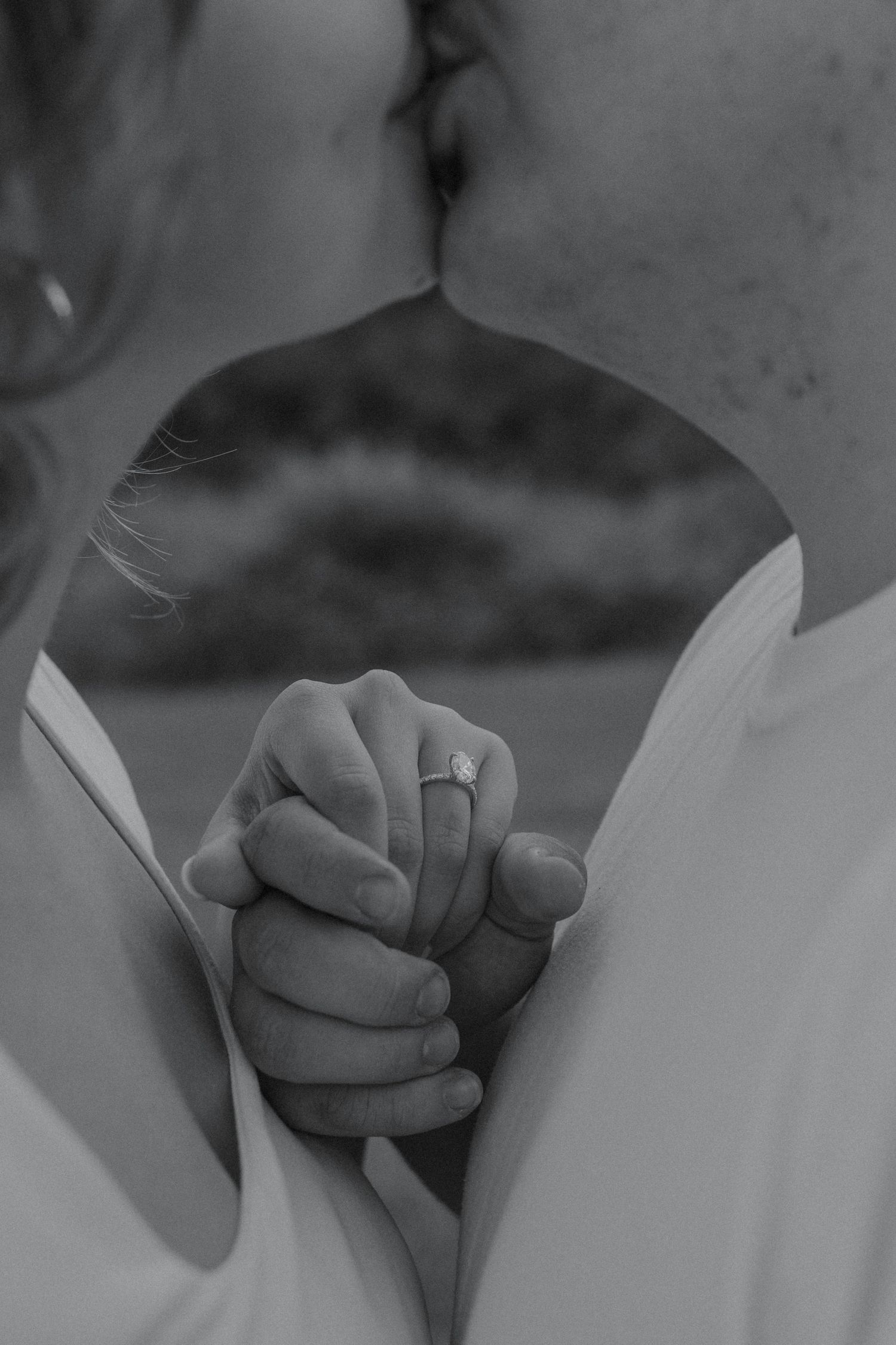 Black and white close-up of engaged couple kissing while holding hands showing diamond engagement ring.