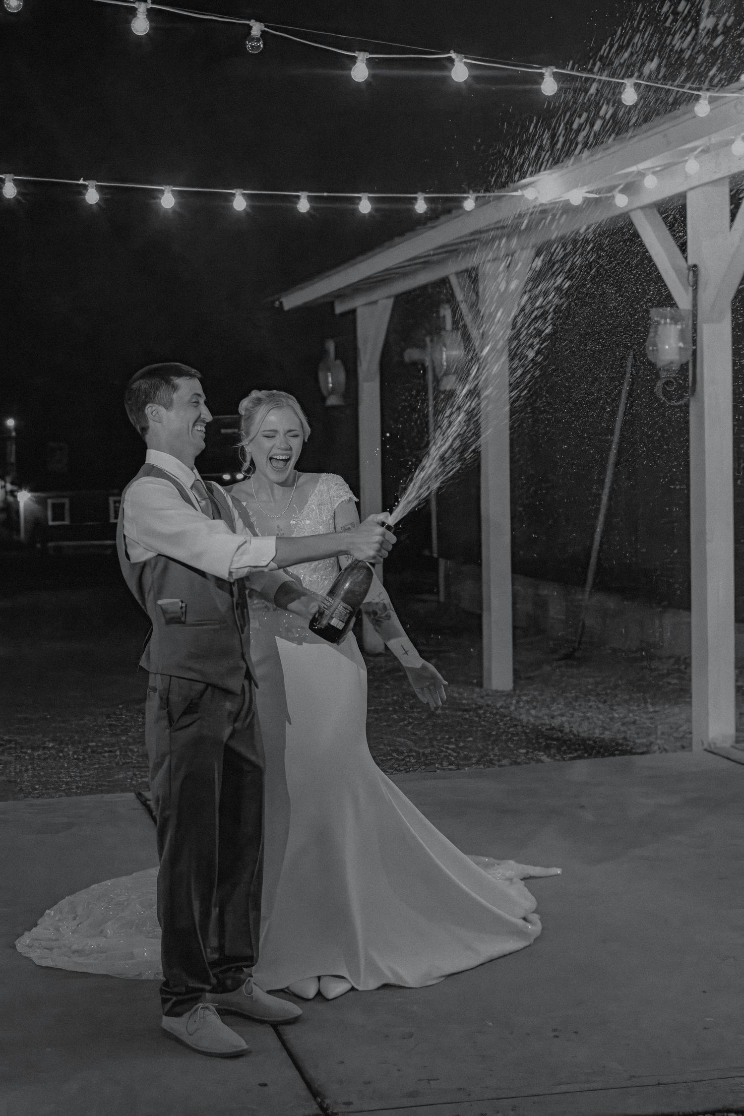 Black and white photo of newlyweds popping champagne bottle outdoors under string lights at night.