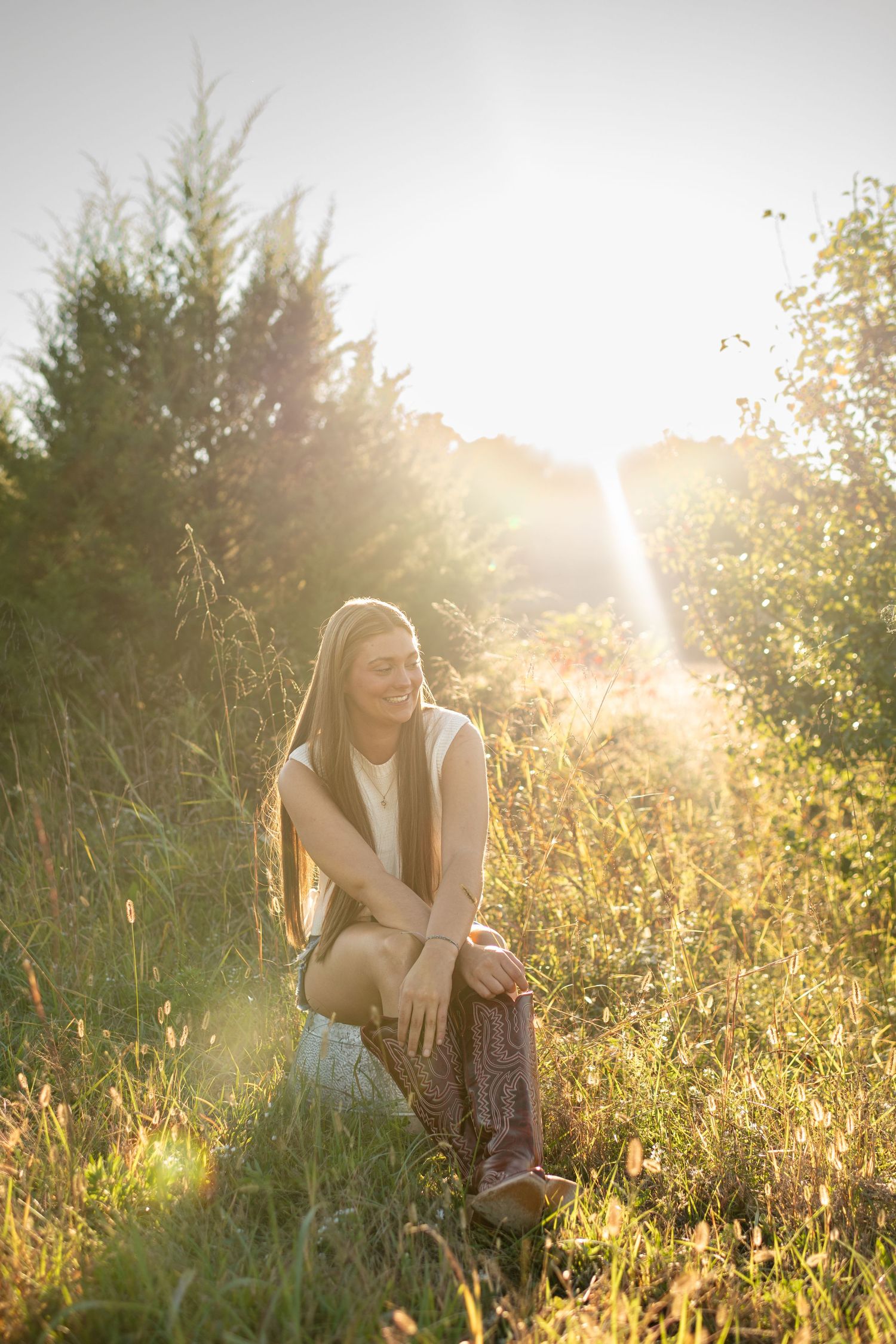 Young woman with long hair smiling while sitting in a sunlit meadow during golden hour, surrounded by tall grass and trees.