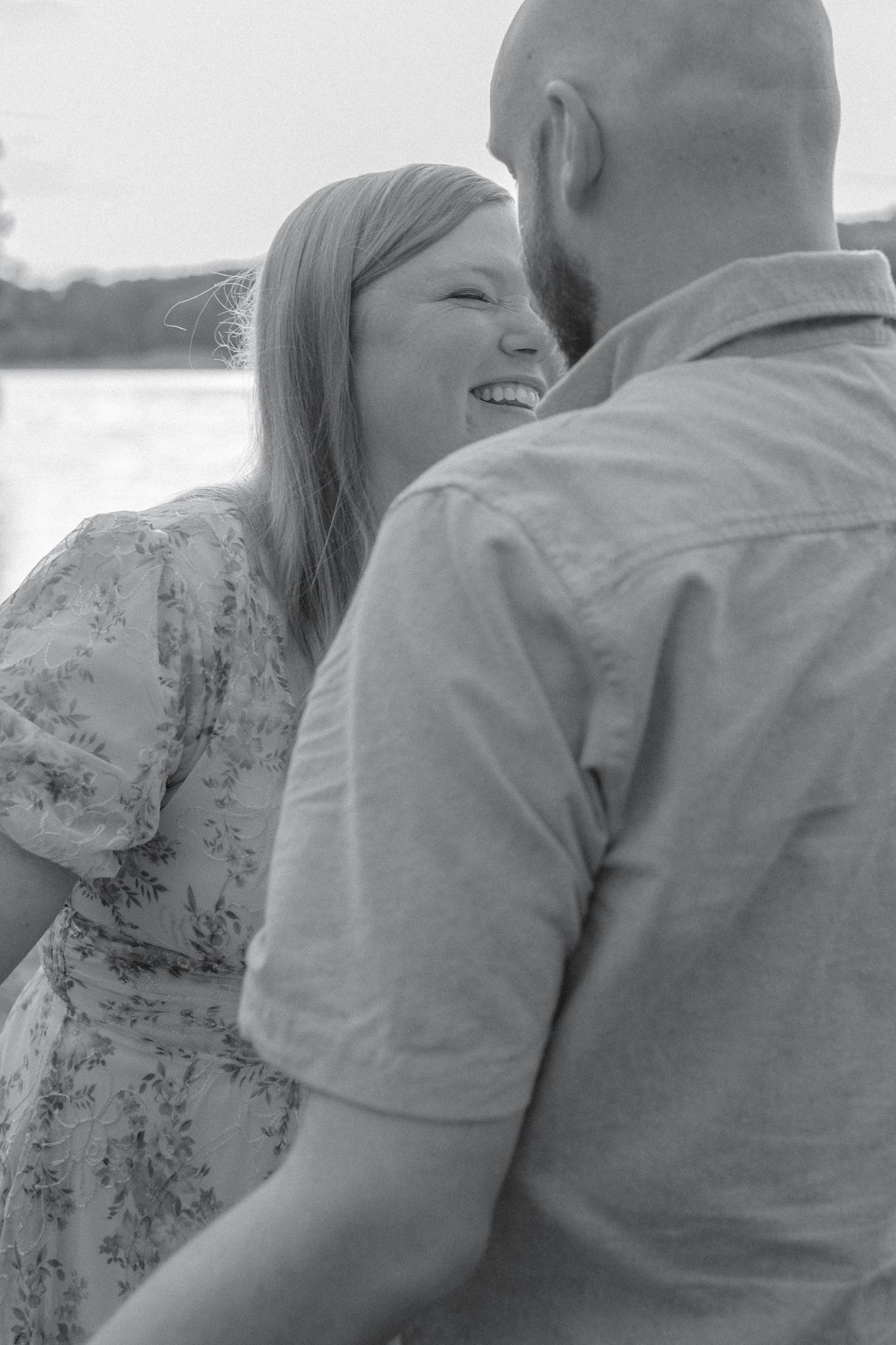 Black and white portrait of smiling woman in floral dress sharing romantic moment with bearded partner outdoors.