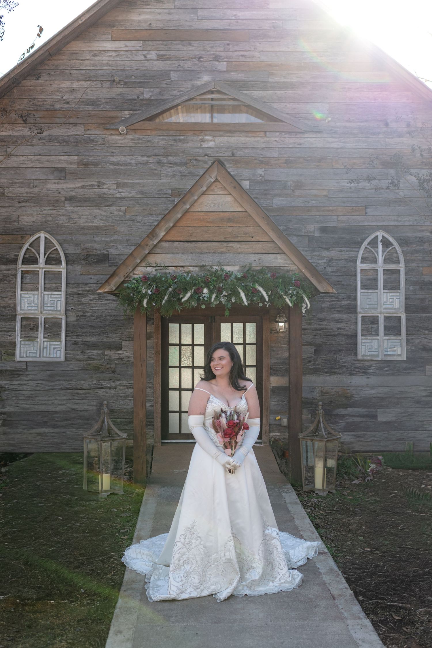 Bride in lace wedding gown stands before a rustic wooden chapel with greenery arch.