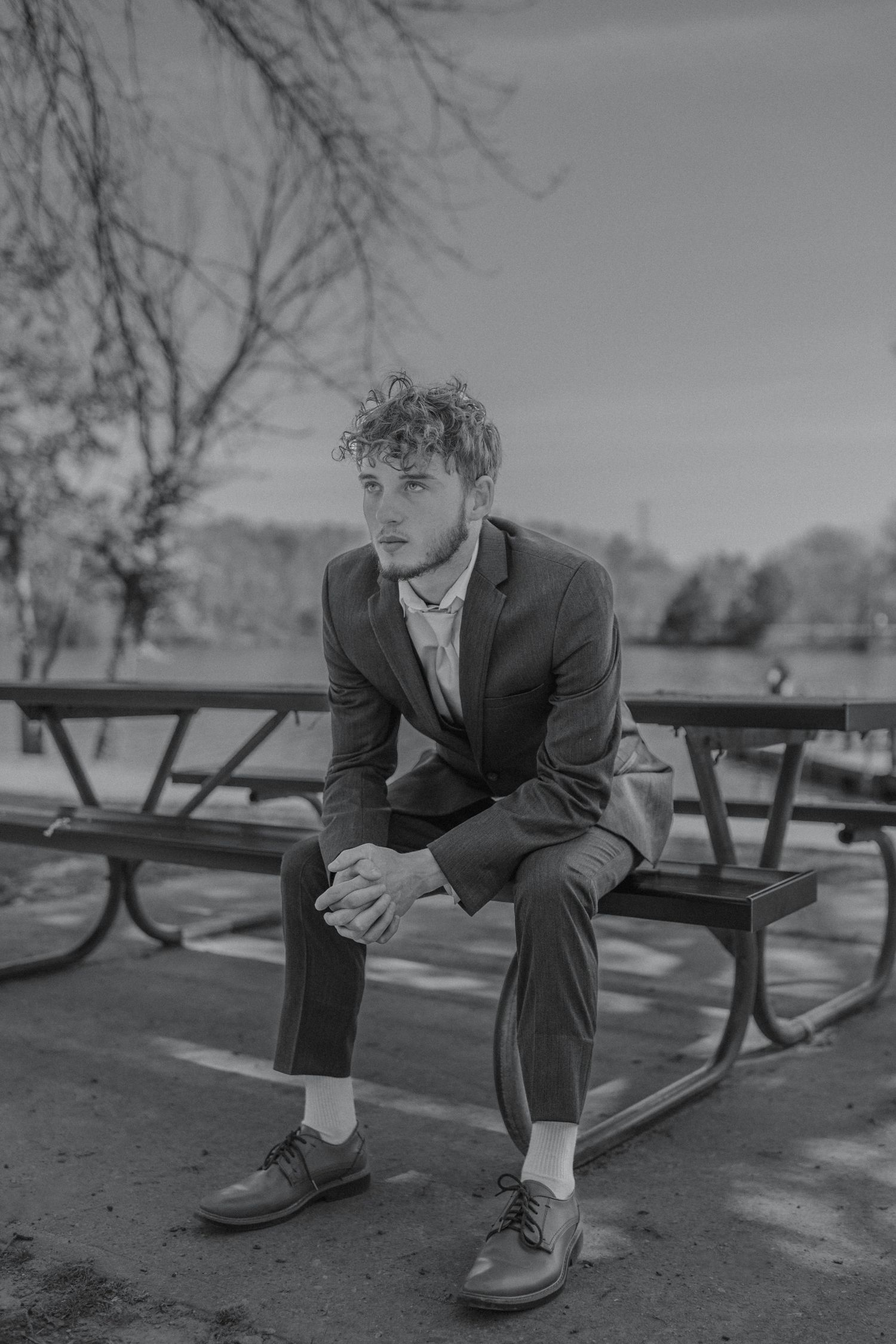 Black and white photo of a young man in a suit sitting on a park bench with hands clasped, looking away.