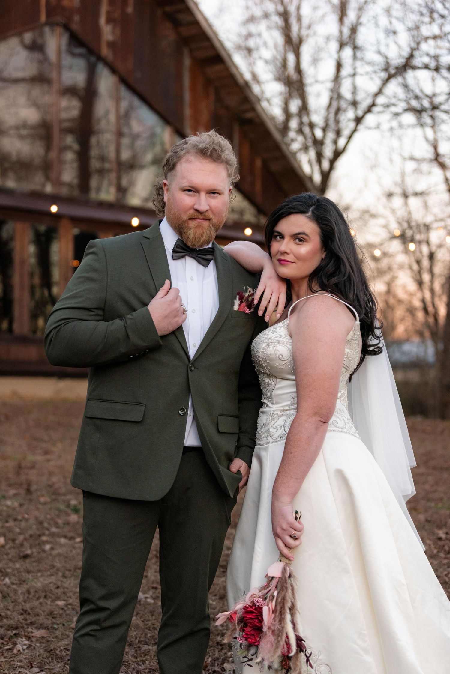 Groom in olive suit and bride in lace gown pose together outside rustic barn venue.