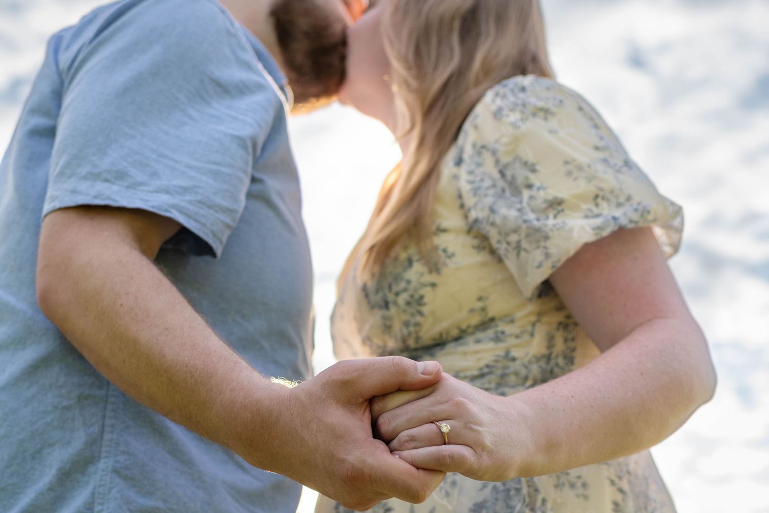Close-up of engaged couple holding hands outdoors, woman wearing diamond ring in floral yellow dress.